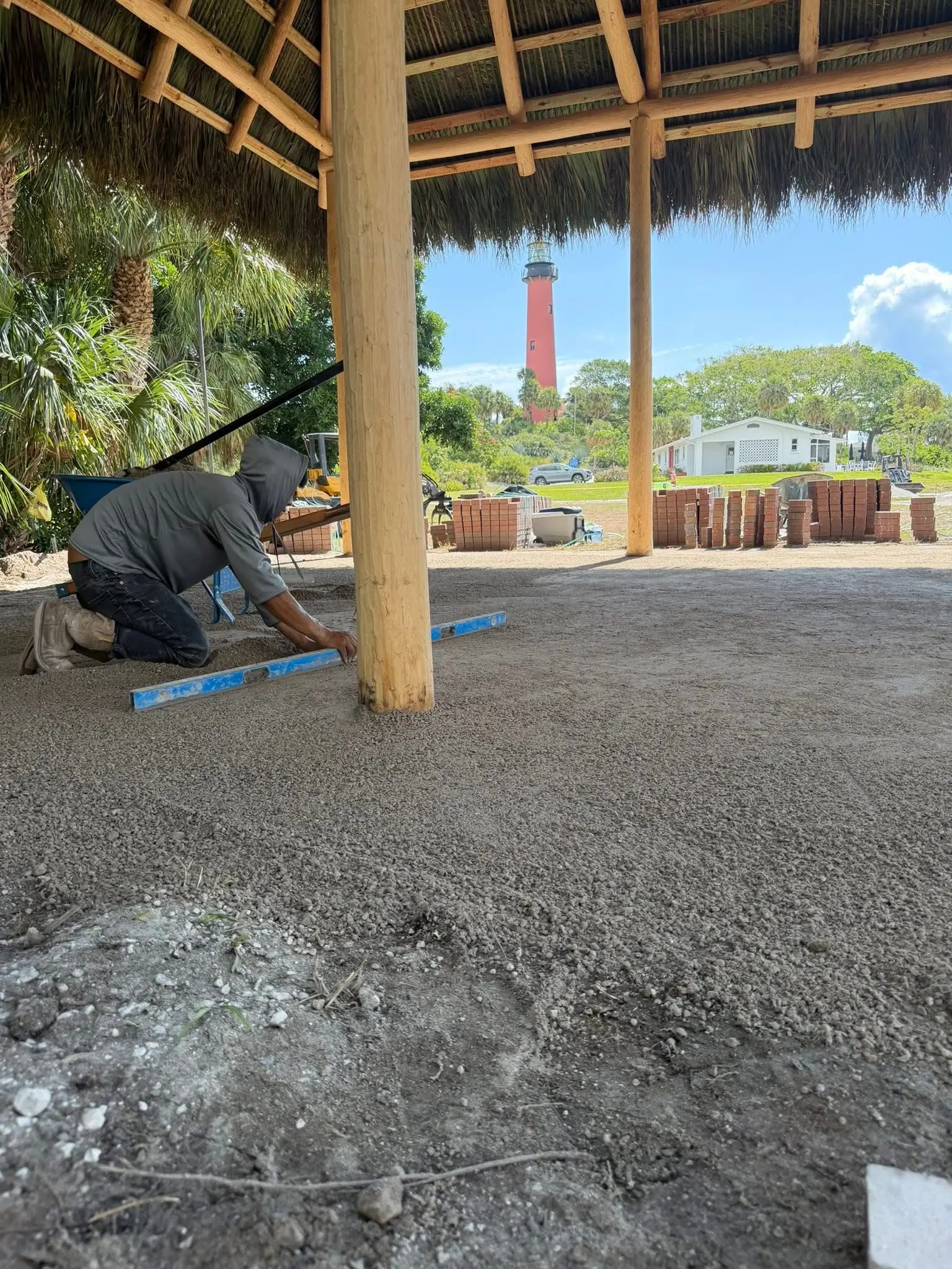 Hardscape paver base preparation by Integrity Landscape showing a worker leveling a sand base under a thatched chickee hut with the Jupiter Lighthouse framed in the background