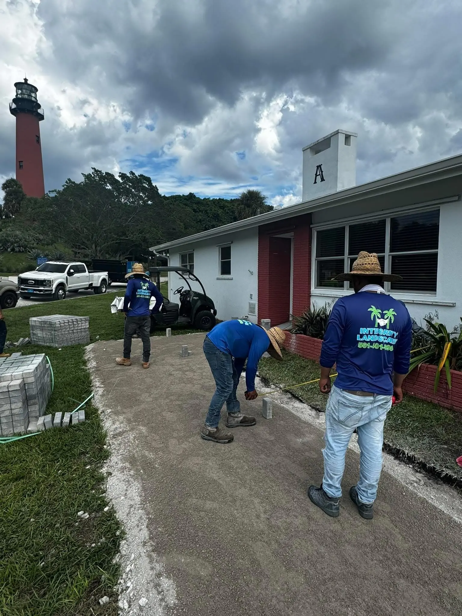 Hardscape paver installation by Integrity Landscape showing three crew members in branded blue shirts placing concrete pavers at the Jupiter Lighthouse grounds