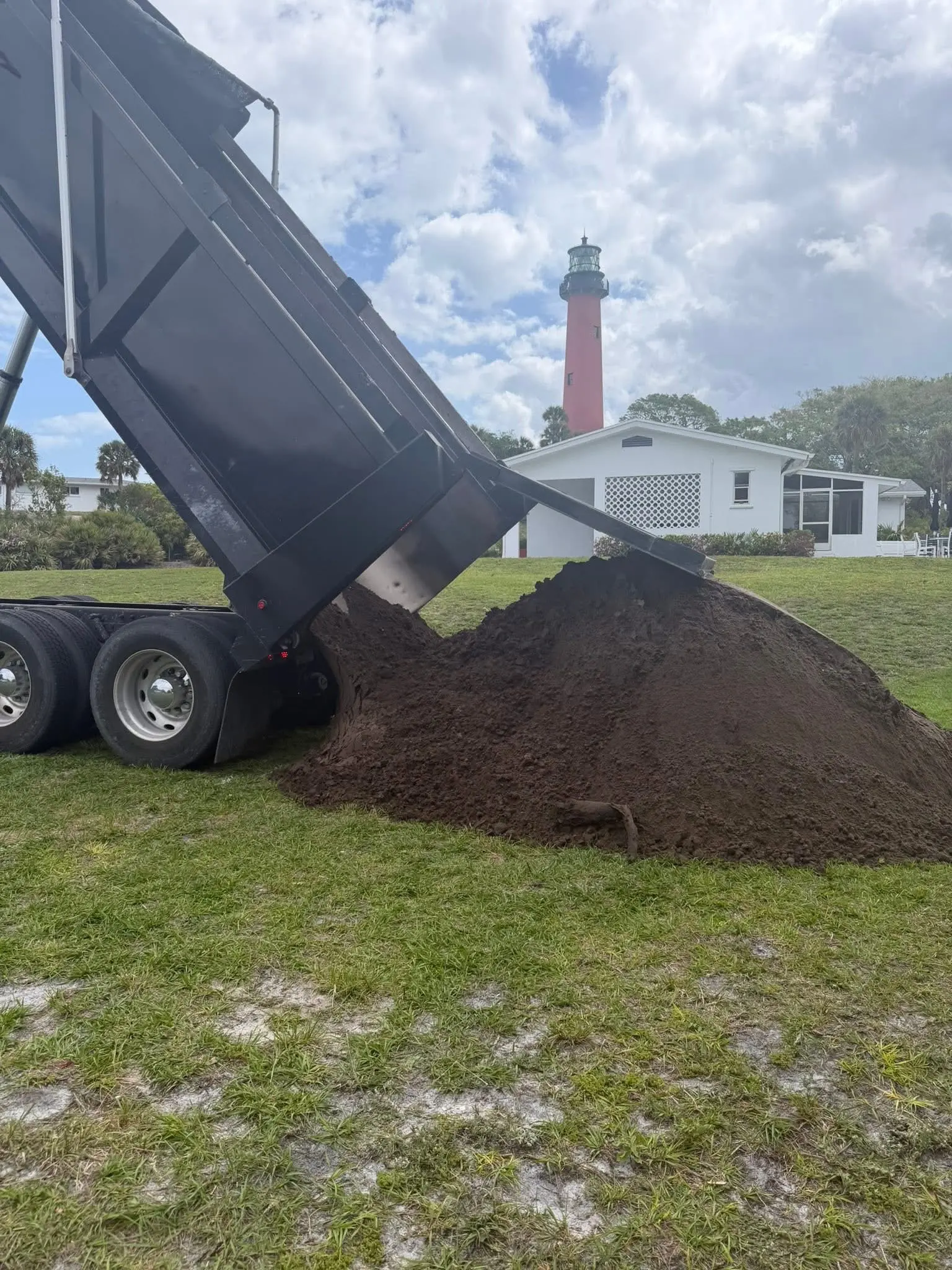 Landscape soil delivery by Integrity Landscape showing a dump truck unloading a large mound of dark rich topsoil onto a residential lawn with the Jupiter Lighthouse visible in the background