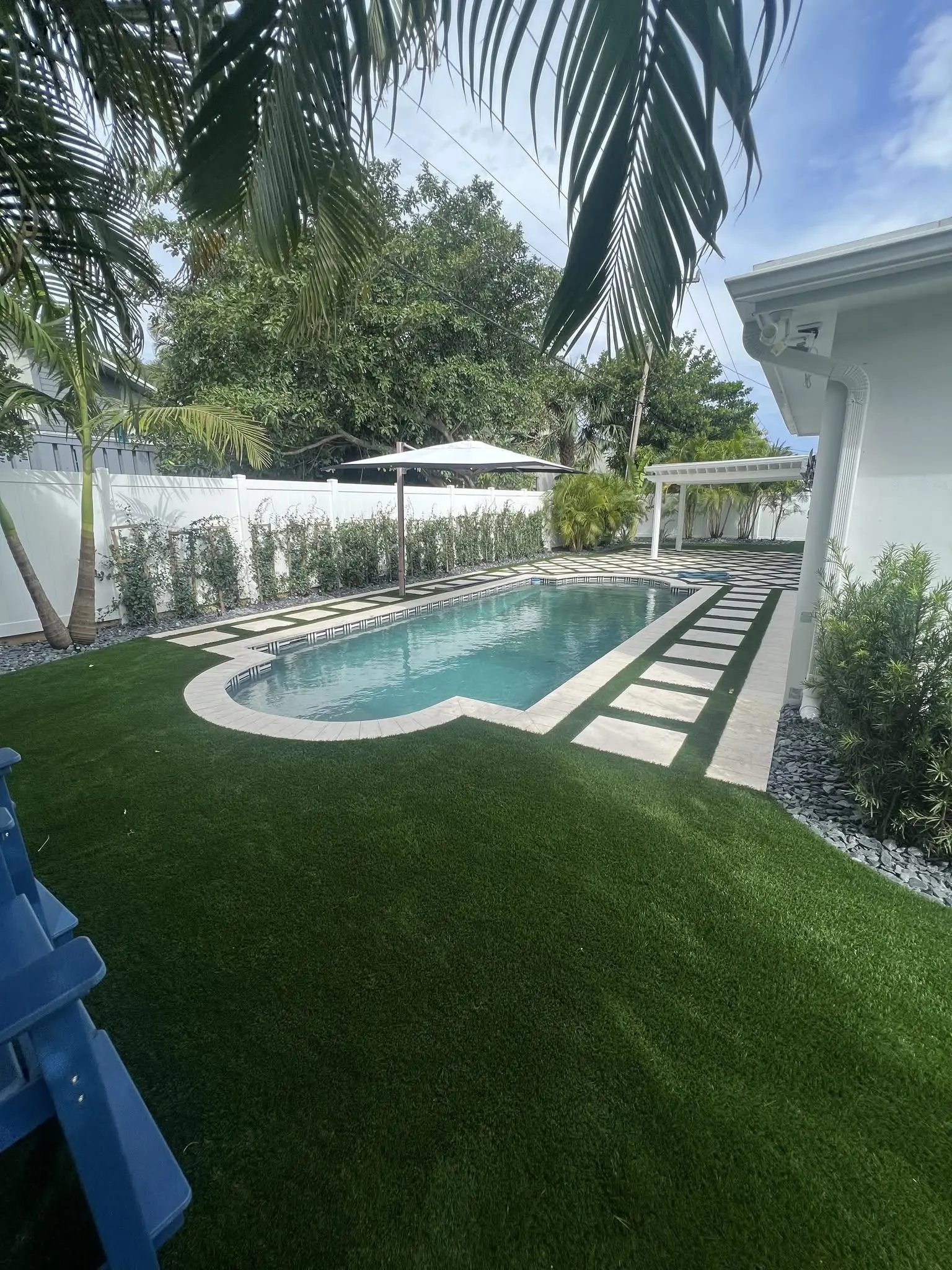 Landscape and hardscape installation by Integrity Landscape showing a completed backyard with pristine artificial turf surrounding a rectangular pool featuring a striking checkerboard black-and-white tile paver deck, white vinyl fence, white pergola, and manicured ficus hedgerow in Palm Beach County