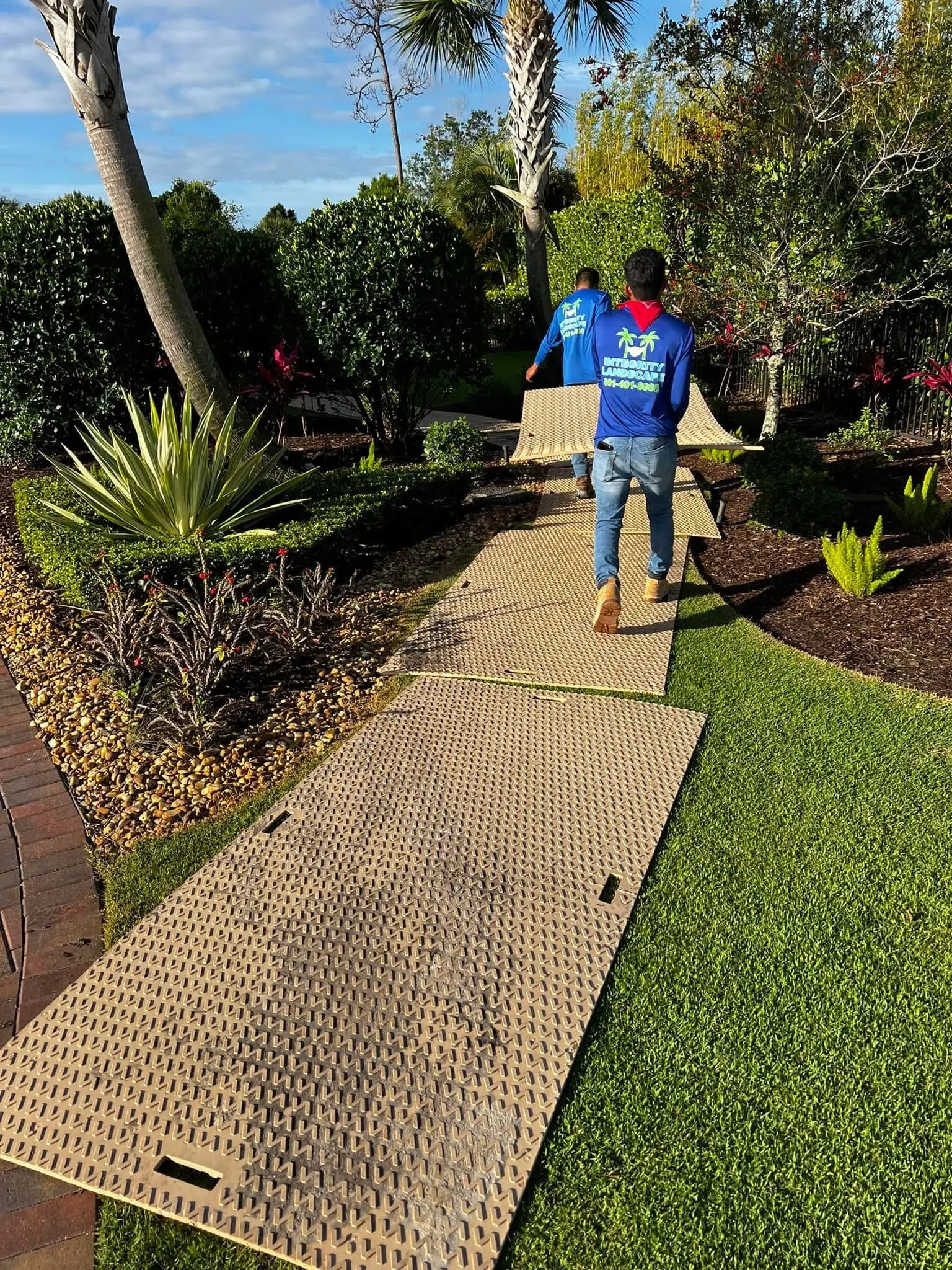 Landscape installation by Integrity Landscape showing two crew members in branded blue shirts walking a freshly completed artificial turf path flanked by colorful tropical garden beds with bromeliads, agave, and mulch borders along a herringbone brick edge in a Palm Beach County backyard