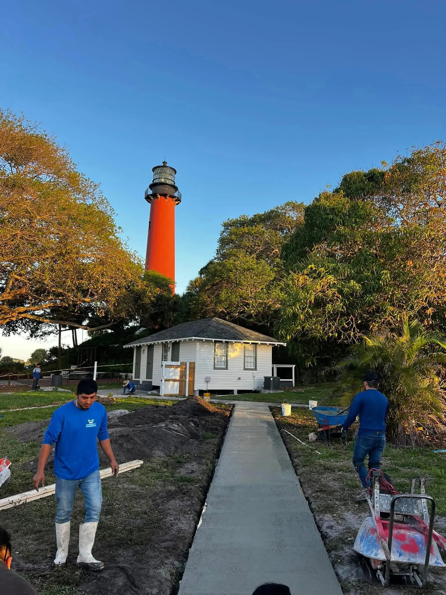 Hardscape concrete walkway installation by Integrity Landscape showing two crew members in branded blue shirts finishing a freshly poured concrete pathway at golden hour, with the iconic Jupiter Lighthouse rising in the background