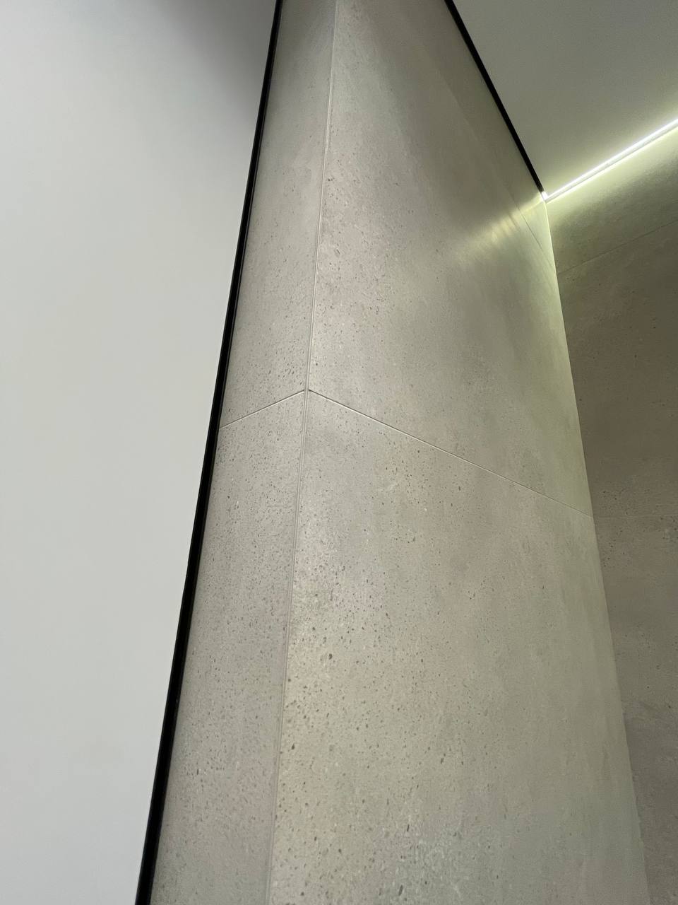 Light concrete tile corner detail inside shower niche with black aluminum trim and warm LED ceiling strip