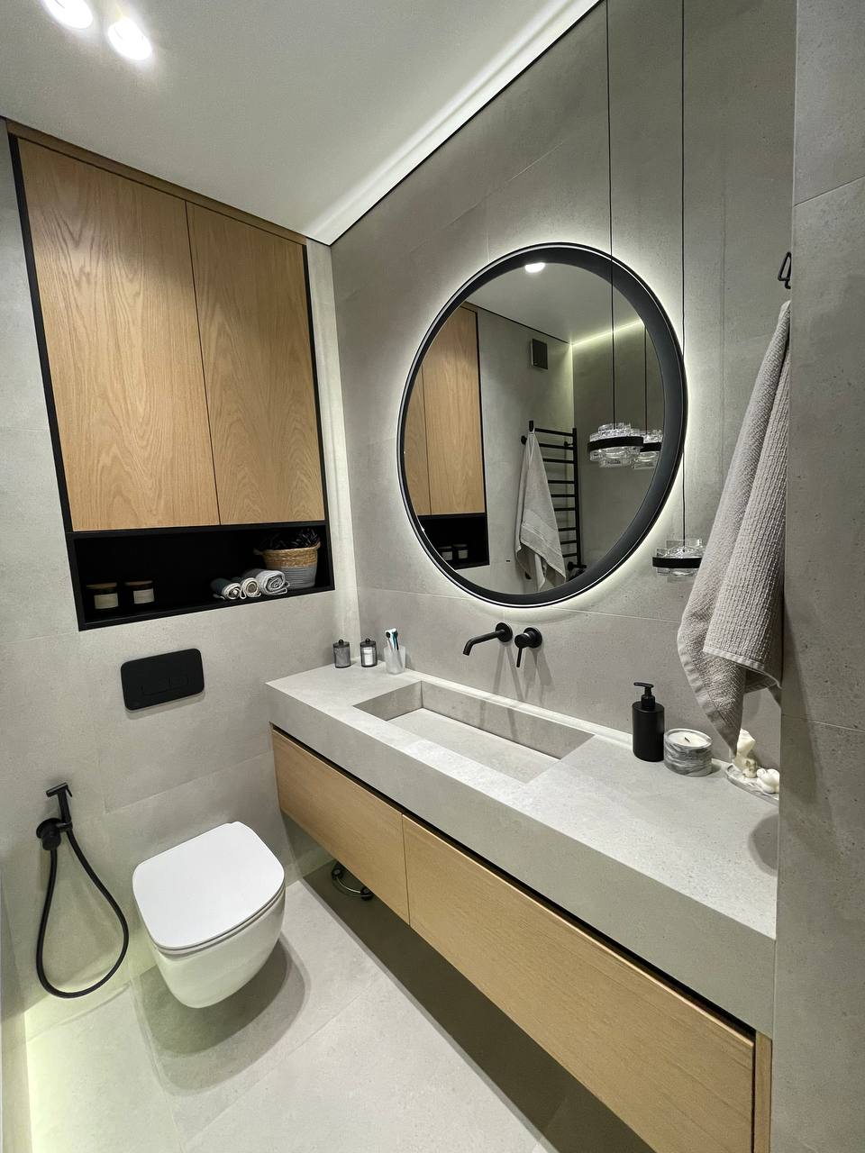Full bathroom with round backlit mirror, oak vanity, integrated concrete sink and crystal pendant lights
