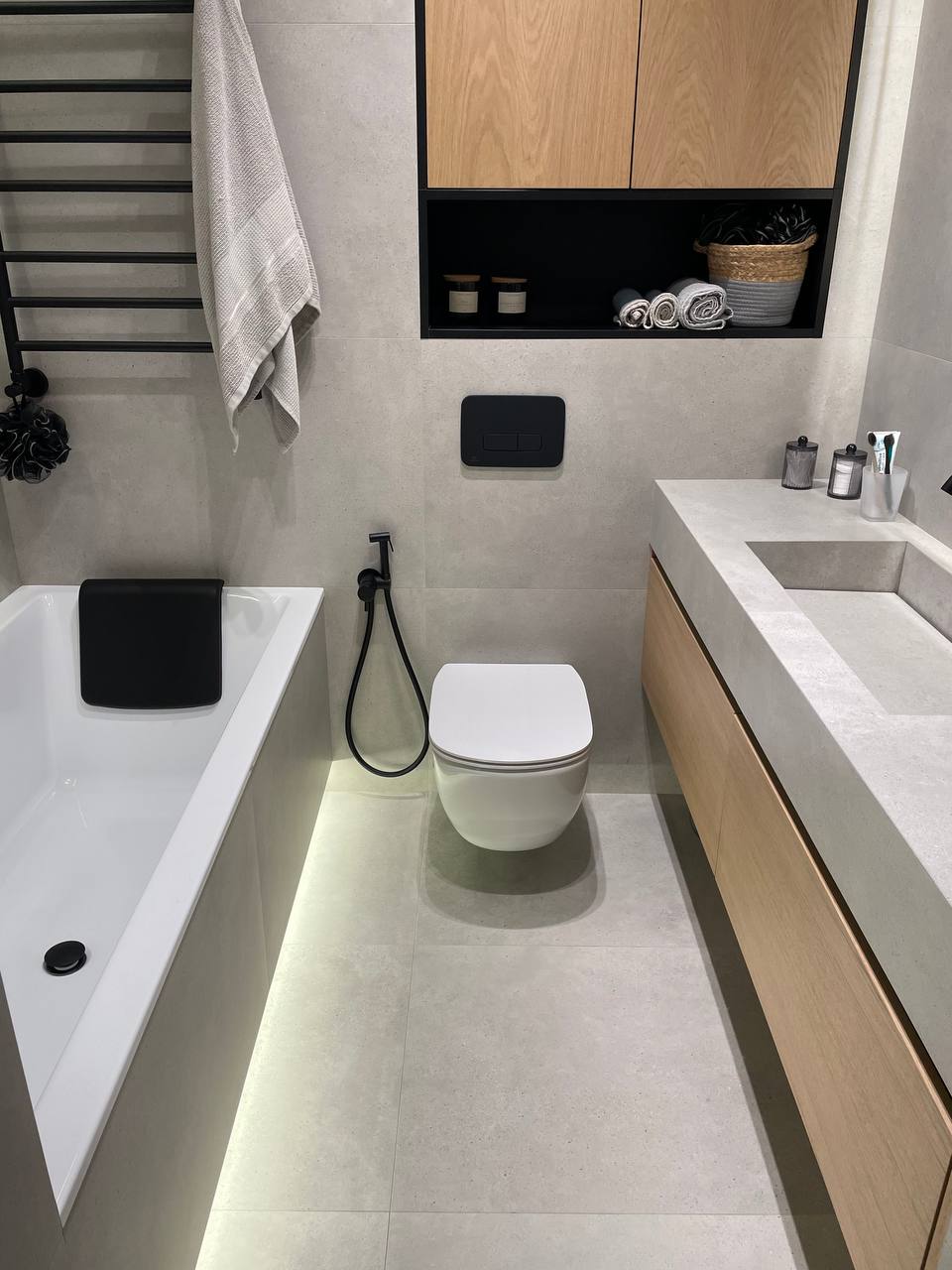 Bathroom overview with white bathtub LED underglow, wall-hung toilet, black heated towel rail and oak upper cabinets