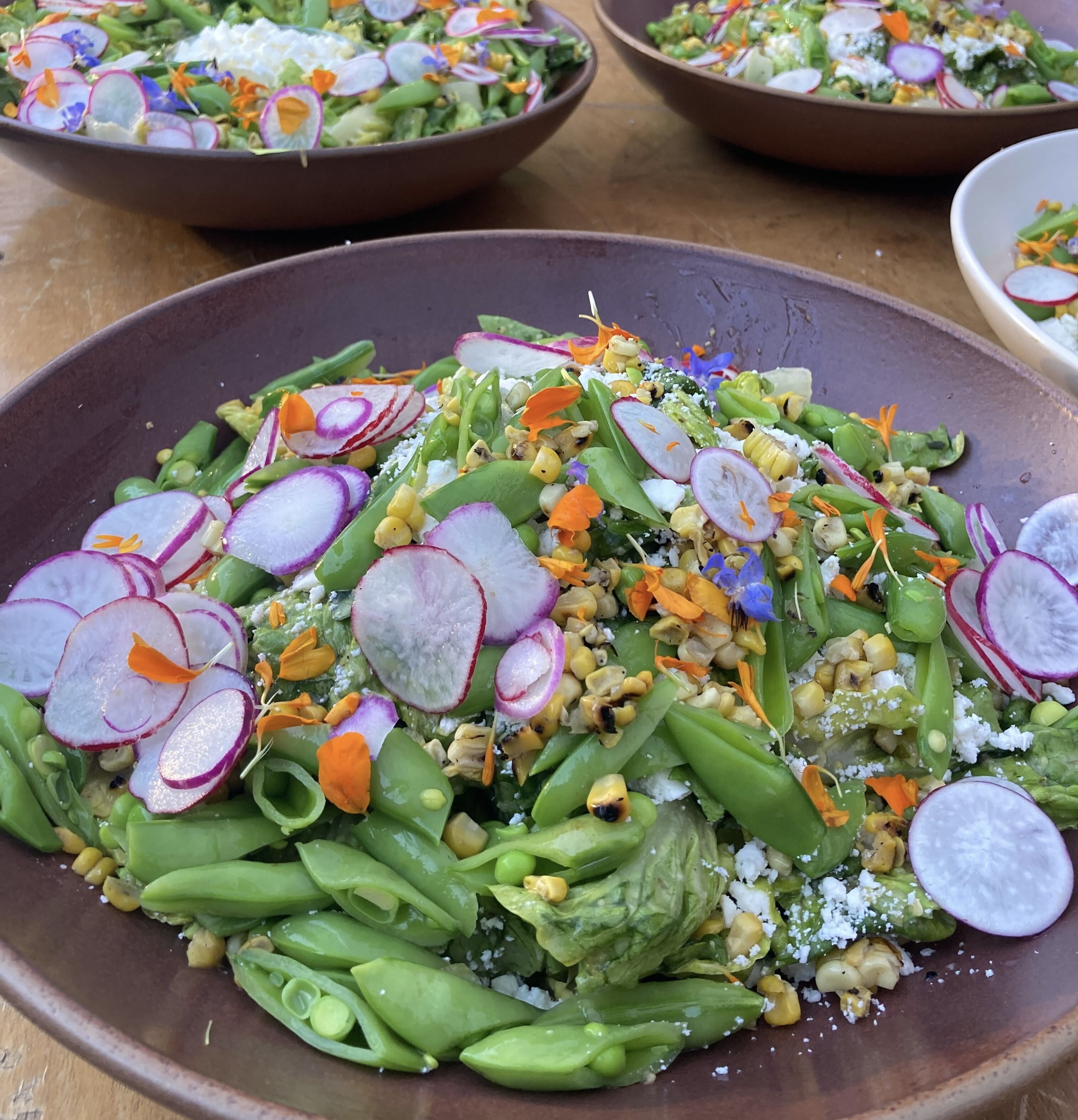 Just Fare workplace event salad with snap peas, grilled corn, thinly sliced radishes, crumbled cotija, and edible flowers in a ceramic bowl