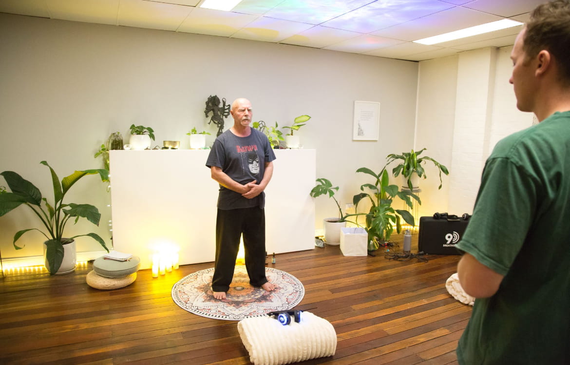 Kadama teaching qigong in his Australian studio, standing barefoot on a mandala rug surrounded by green plants, candles and wooden floors, with a student observing