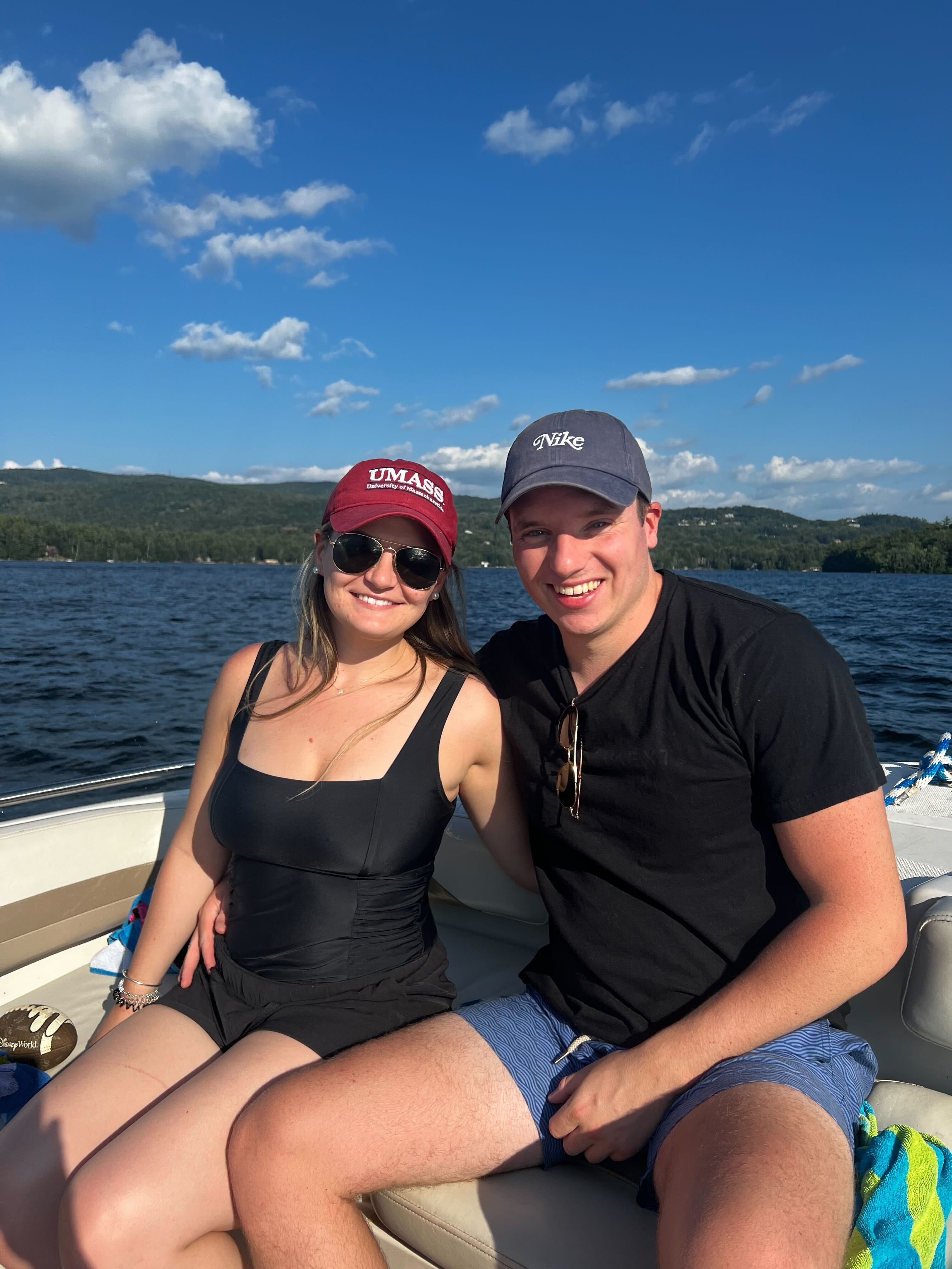 Ben and Brooke enjoying a summer day on a boat, Brooke wearing a UMass cap and Ben in a Nike cap, with a beautiful lake and mountains in the background