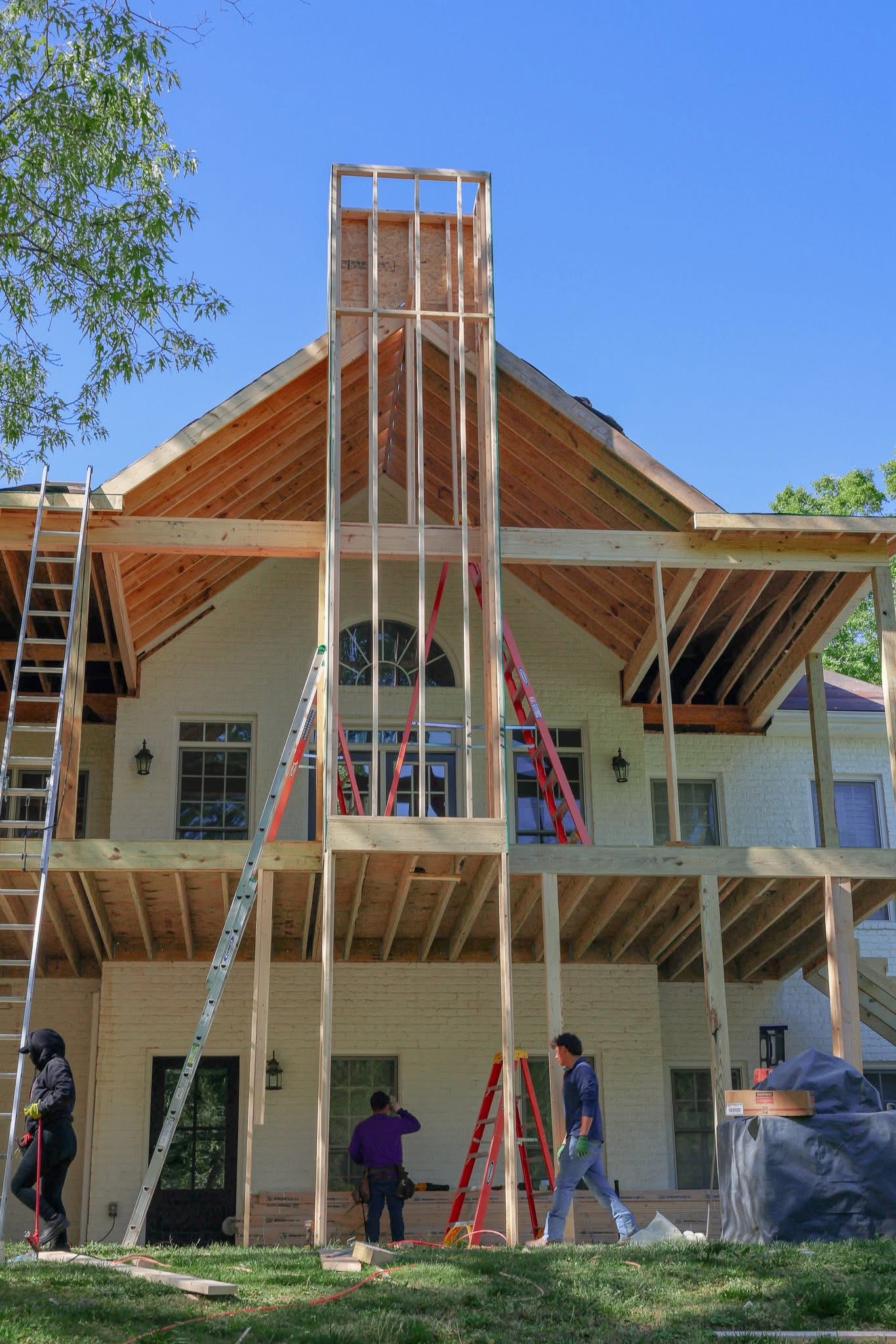 Two-story porch addition mid-framing with fresh lumber structure and crew on site behind a white brick home