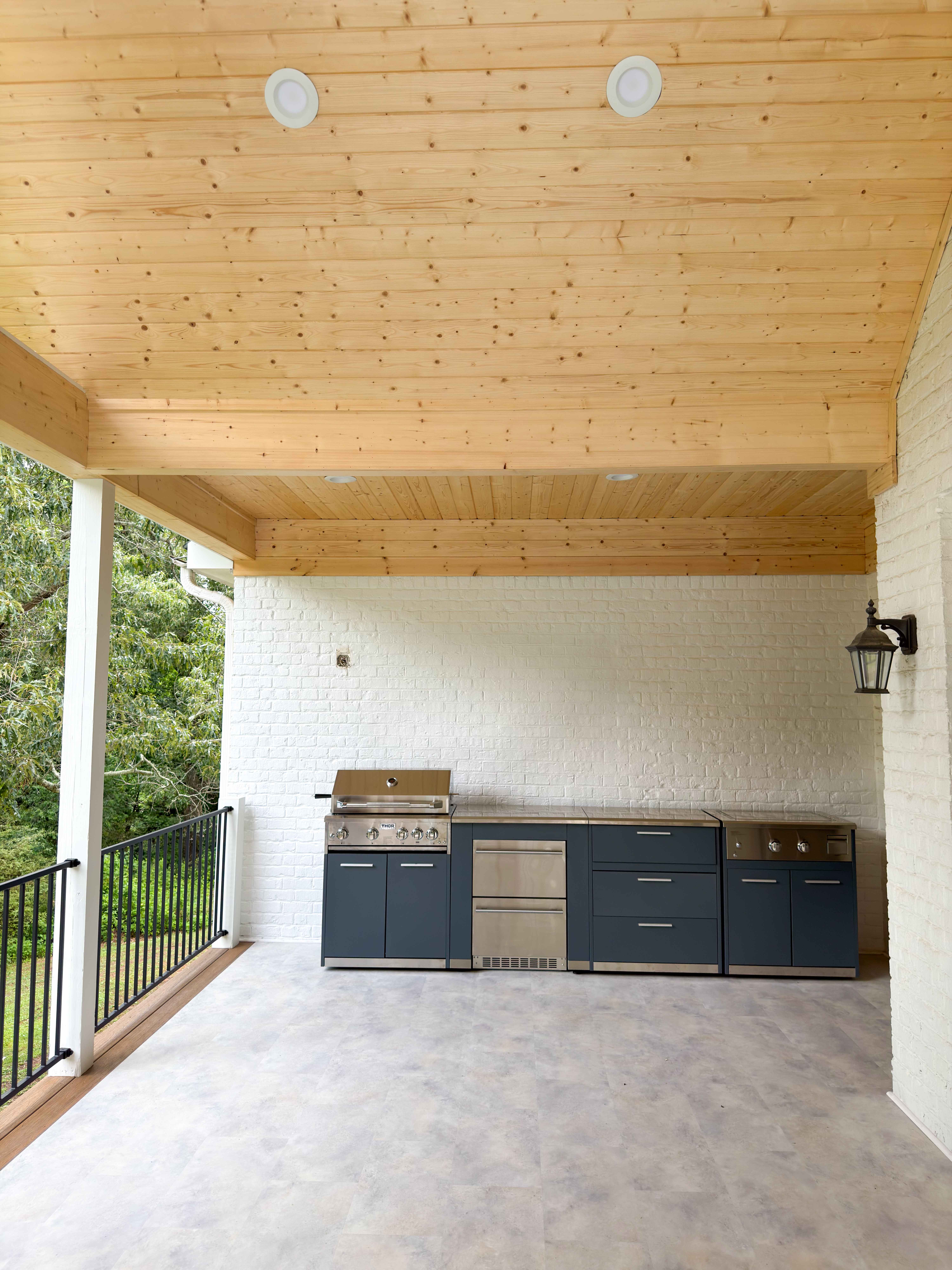 Covered porch outdoor kitchen with a natural-pine plank ceiling, navy cabinetry, a stainless grill and side burner against white painted brick, and a black railing overlooking the trees