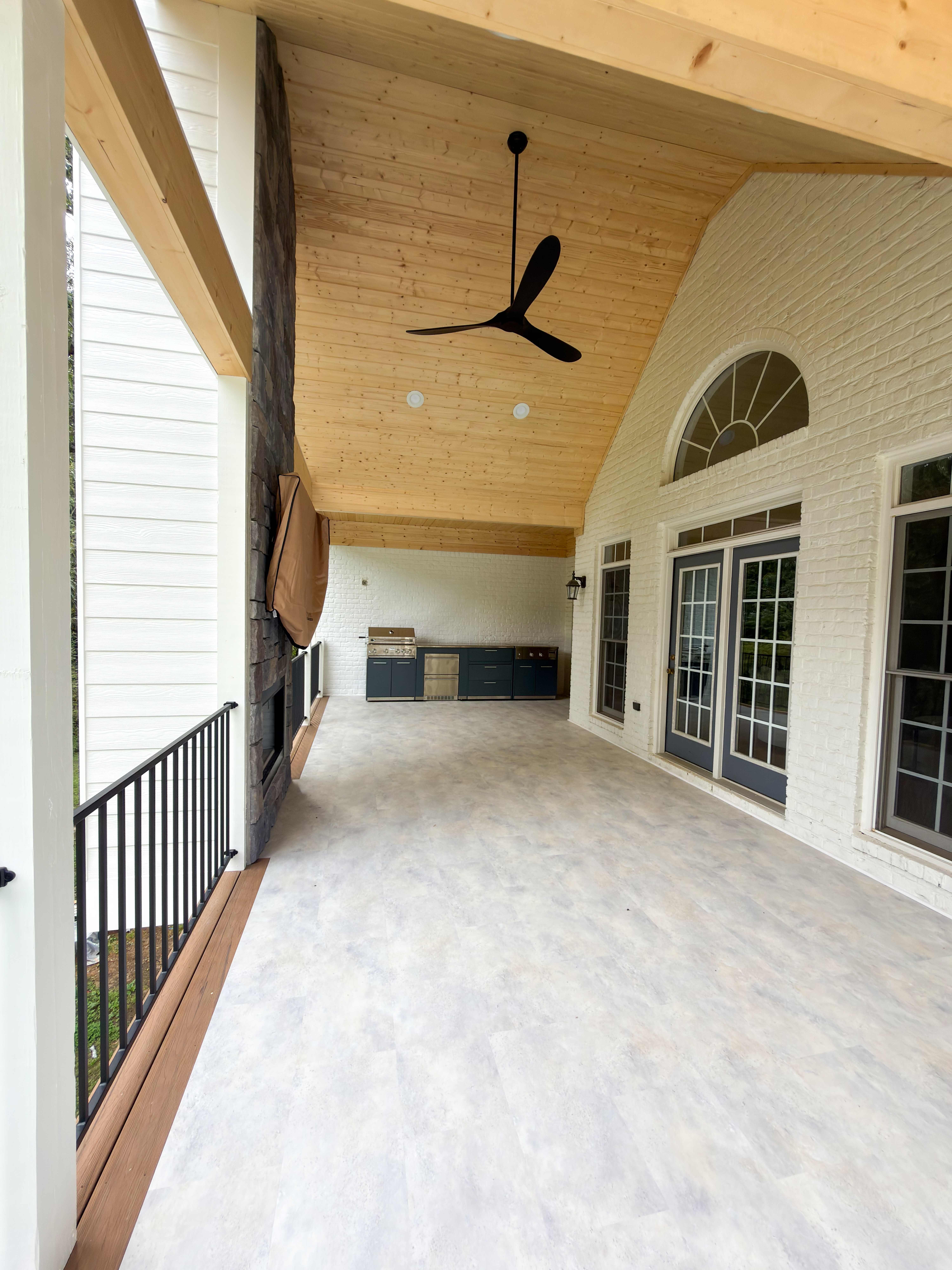 Covered porch with a vaulted natural-pine tongue-and-groove ceiling, a large black modern fan, a stacked-stone fireplace, and a navy outdoor kitchen against white painted brick with arched windows and French doors