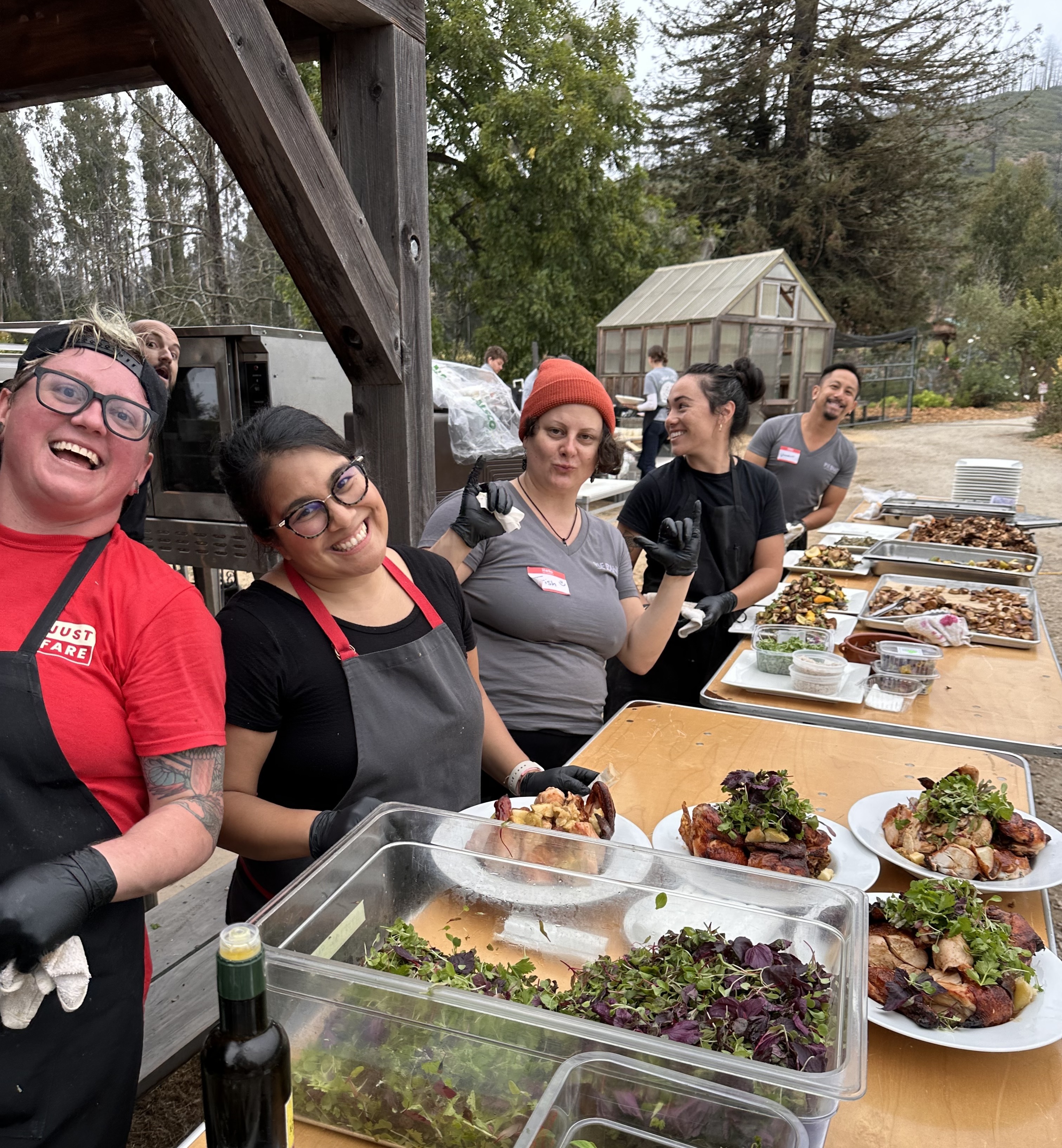 Smiling Just Fare kitchen team in aprons plating roasted chicken and salads at an outdoor event