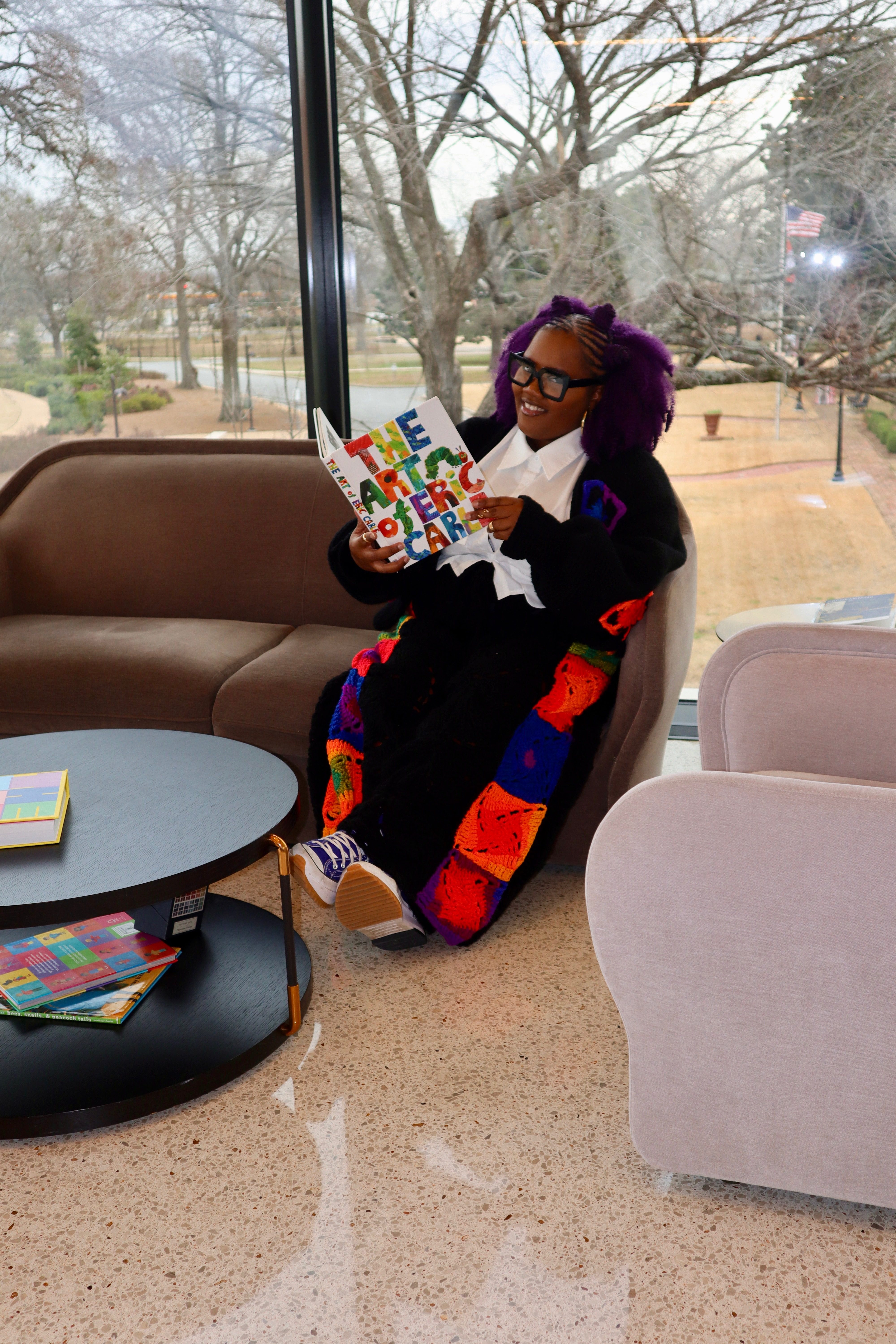Ms. Callie, a passionate tutor with purple hair and glasses, reading The Art of Eric Carle in a cozy library setting