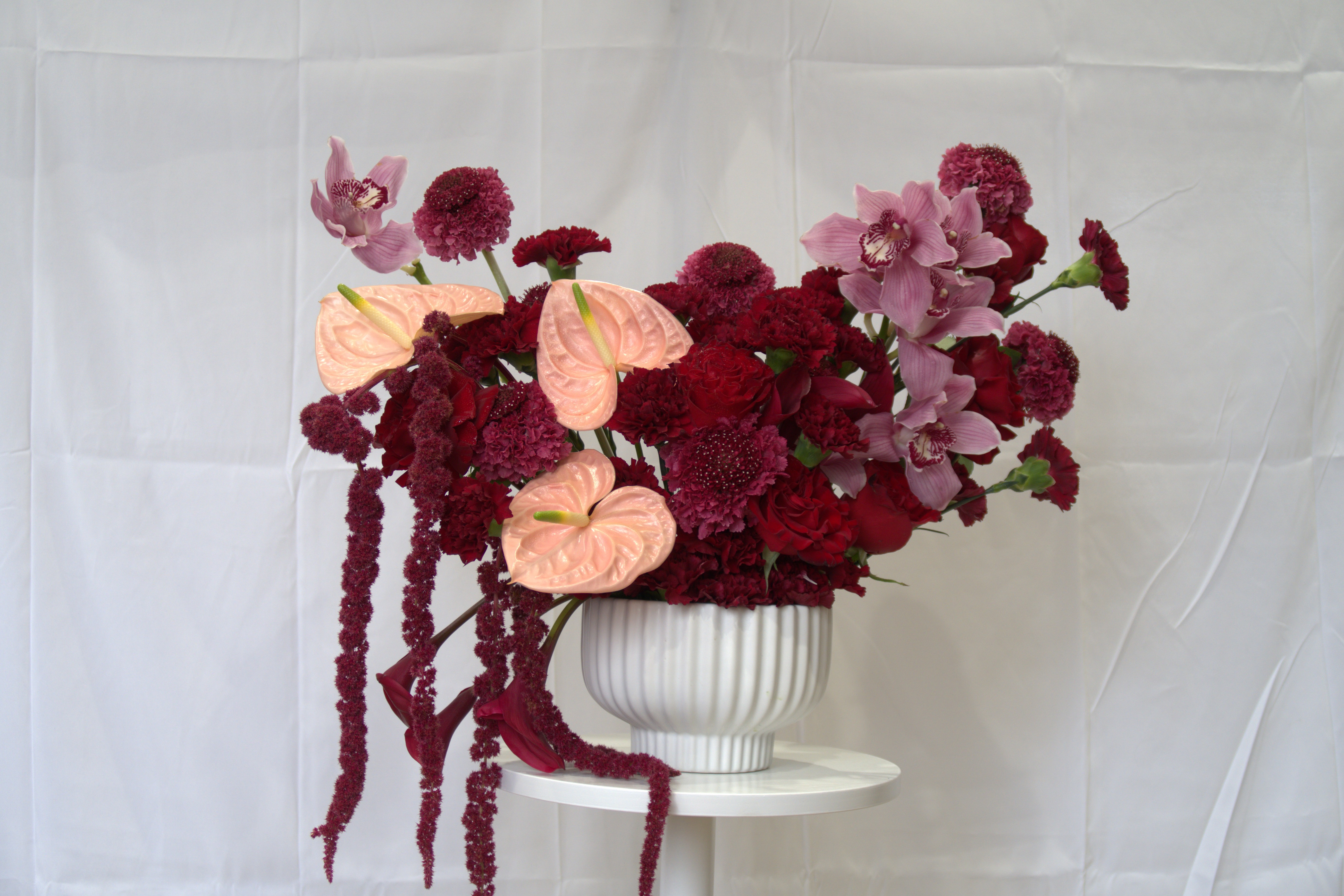 Luxury floral arrangement with burgundy carnations, pink orchids, peach anthuriums and trailing amaranthus in white ribbed bowl