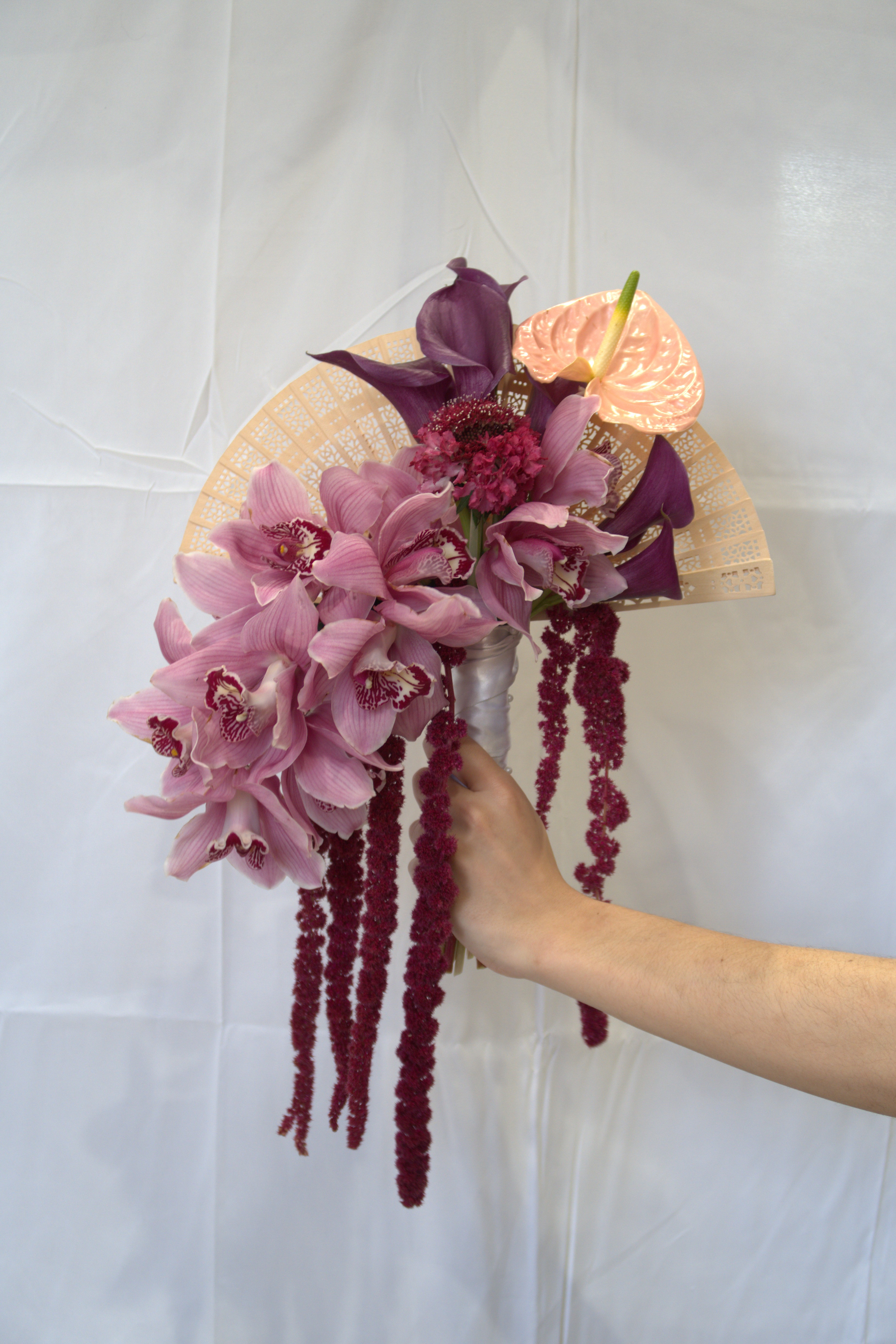 Hand-held bouquet of pink cymbidium orchids, purple calla lilies, peach anthurium and trailing burgundy amaranthus with gold lace fan