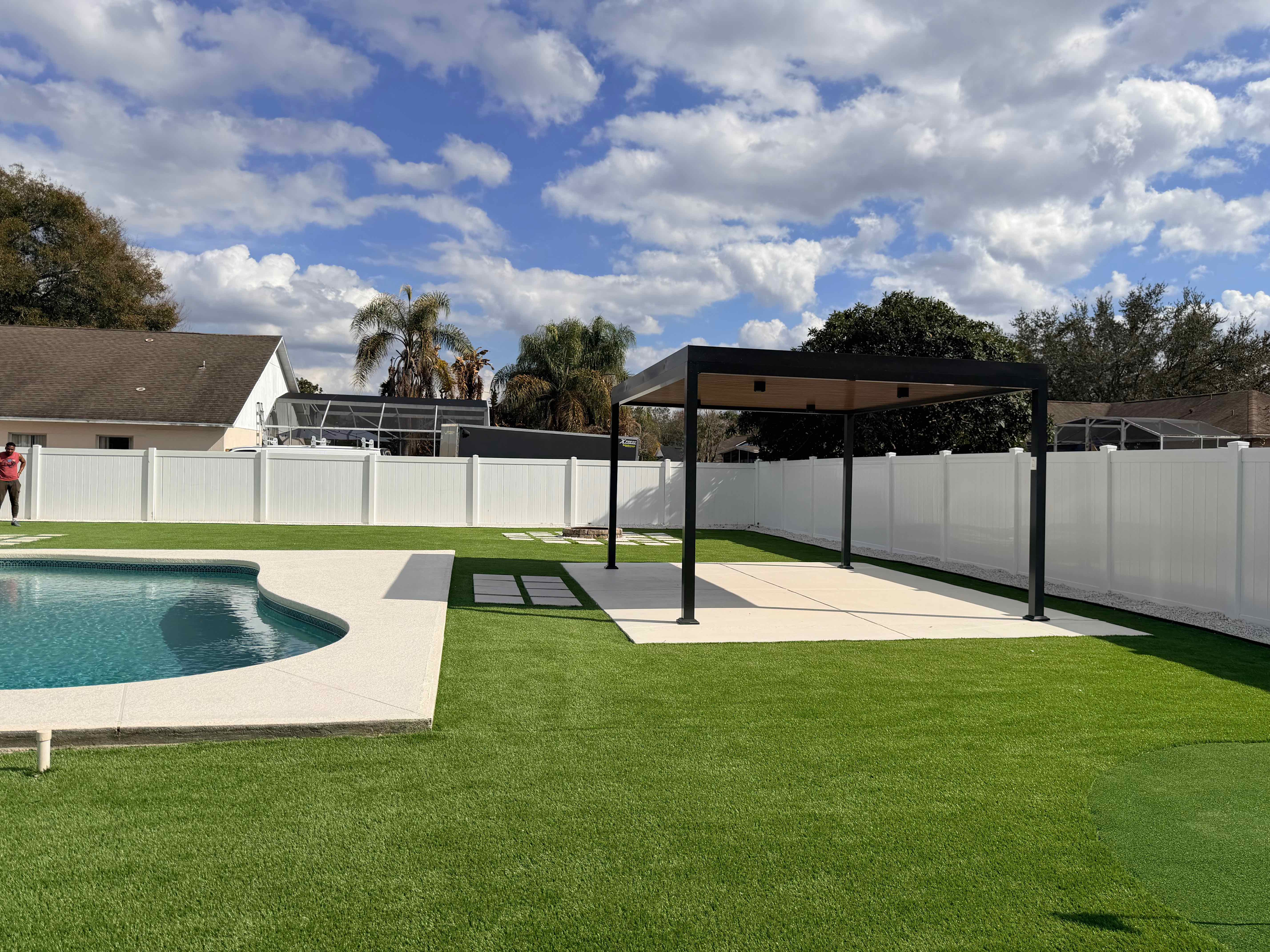 Wide backyard artificial grass installation with pool, black pergola, white vinyl fence and concrete paver strips