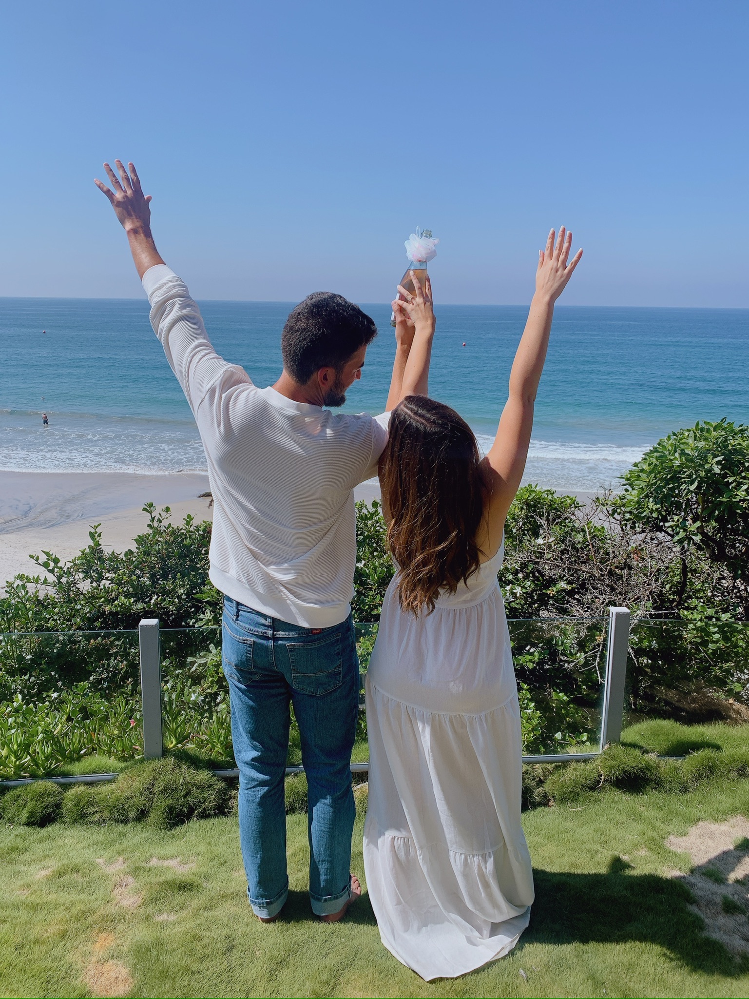 Couple celebrating engagement with arms raised on coastal cliff overlooking ocean