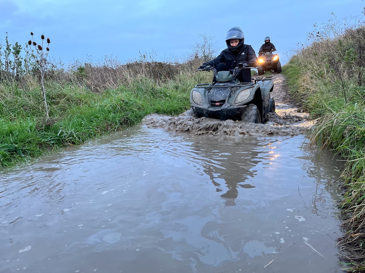 Rider splashing through a deep muddy puddle at dusk with another quad following behind