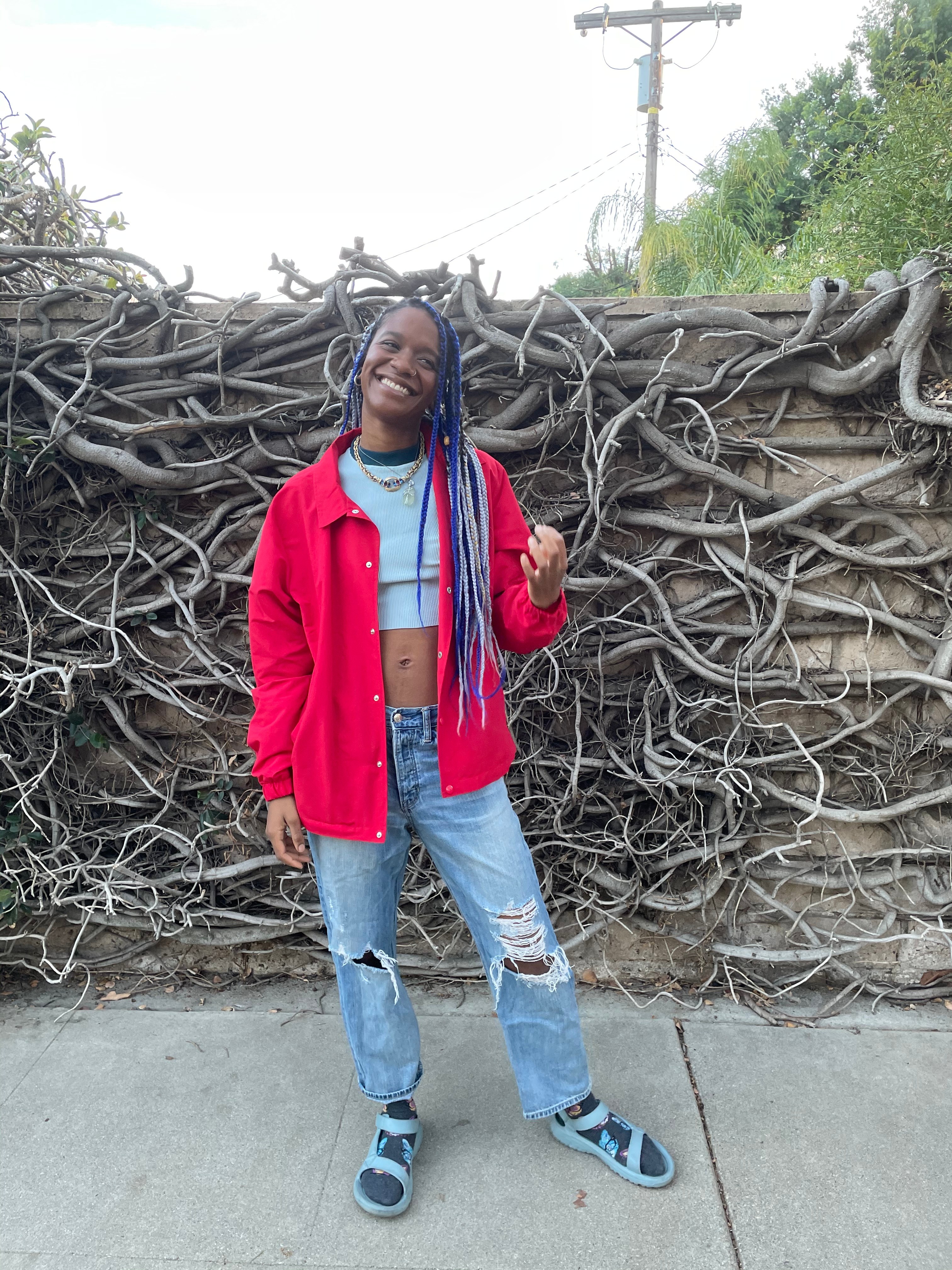 Full-body outdoor shot in red jacket and blue braids against vine wall
