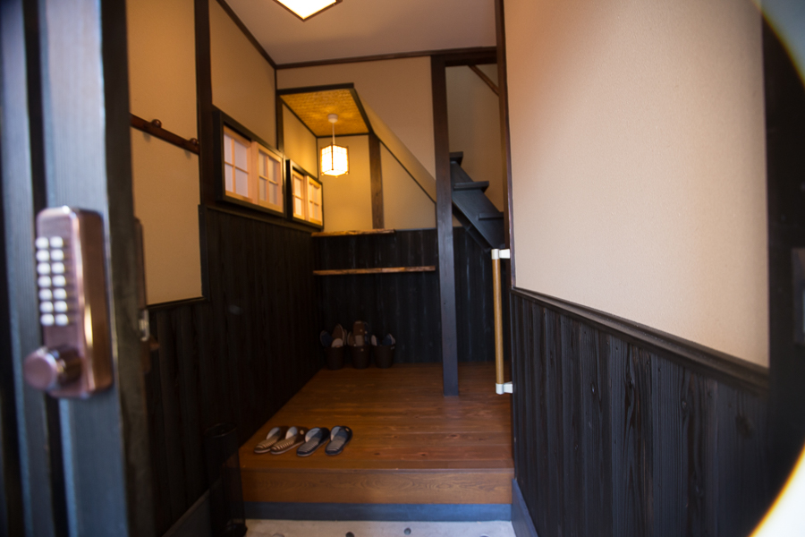 Traditional machiya entrance genkan with dark wood paneling, wooden step, slippers, warm pendant lighting, and keypad lock