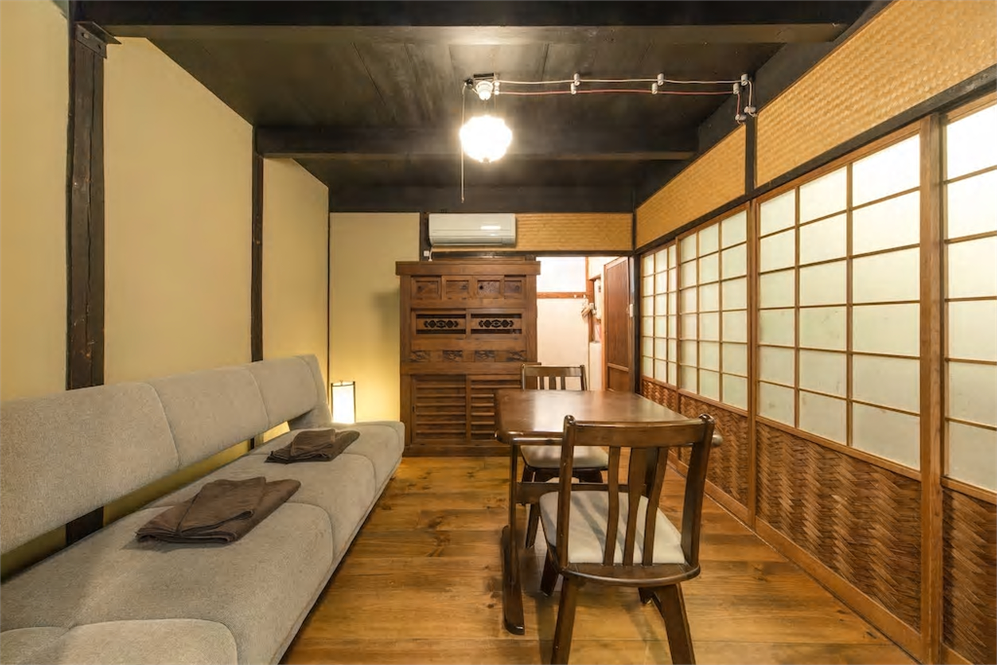 Iho-An machiya living room with long grey sofa, antique tansu cabinet, wooden dining table, shoji screens with woven bamboo ajiro panels, and warm paper lantern