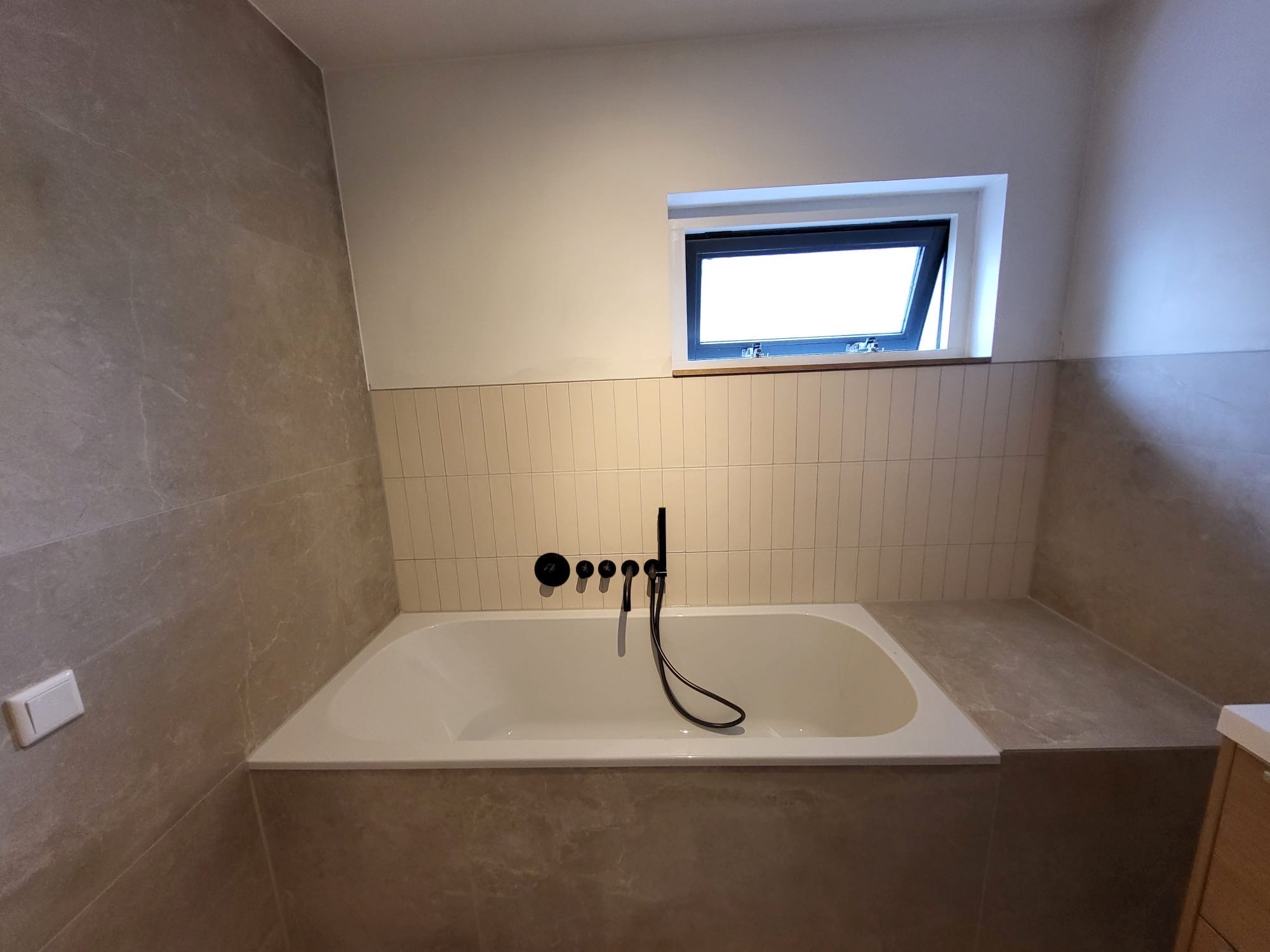 Ingebouwd ligbad met zwarte inbouwkranen tegen een wand van crèmekleurige staande tegels