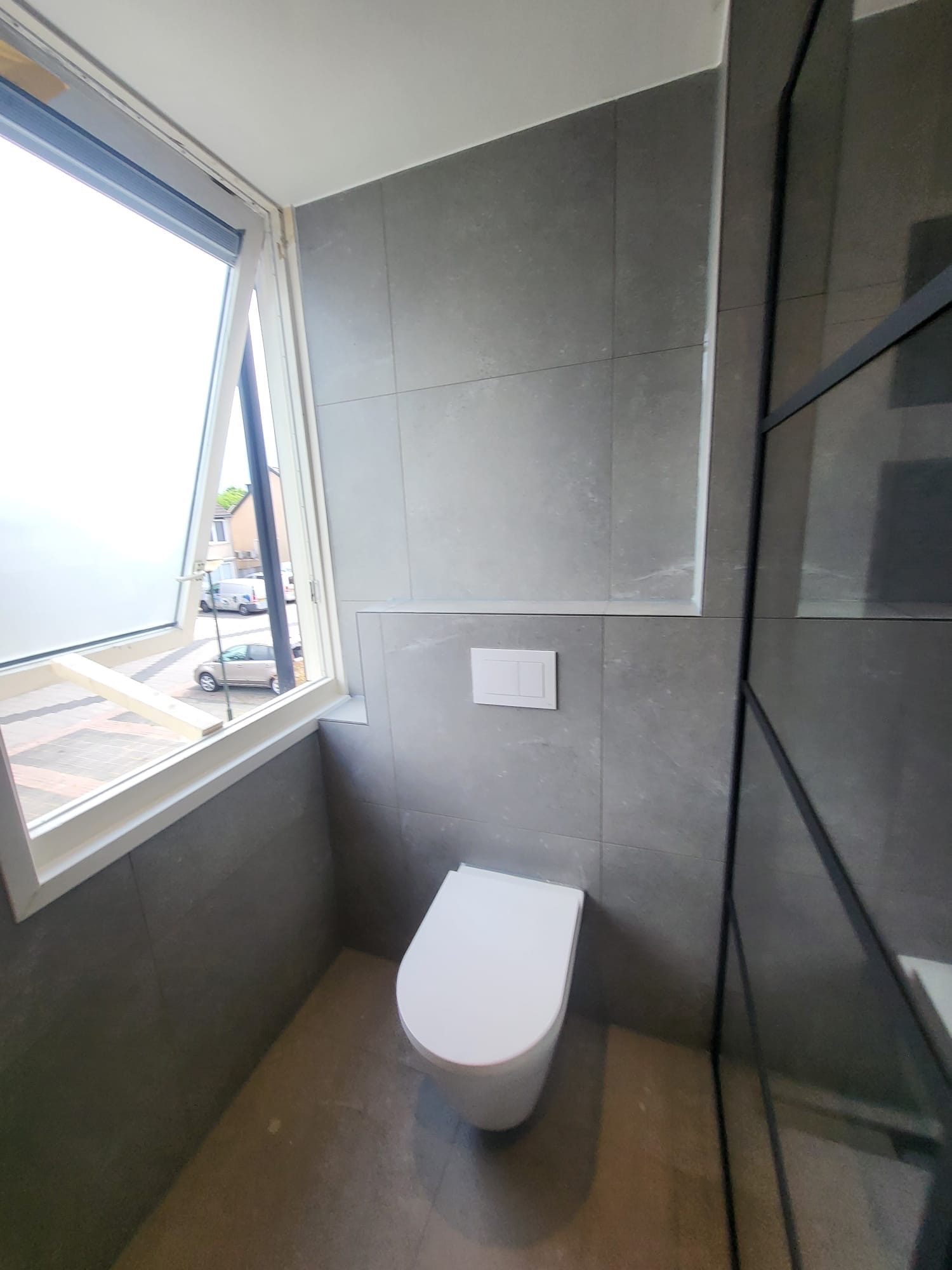 Hangtoilet tegen een wand met grote grijze tegels naast een raam
