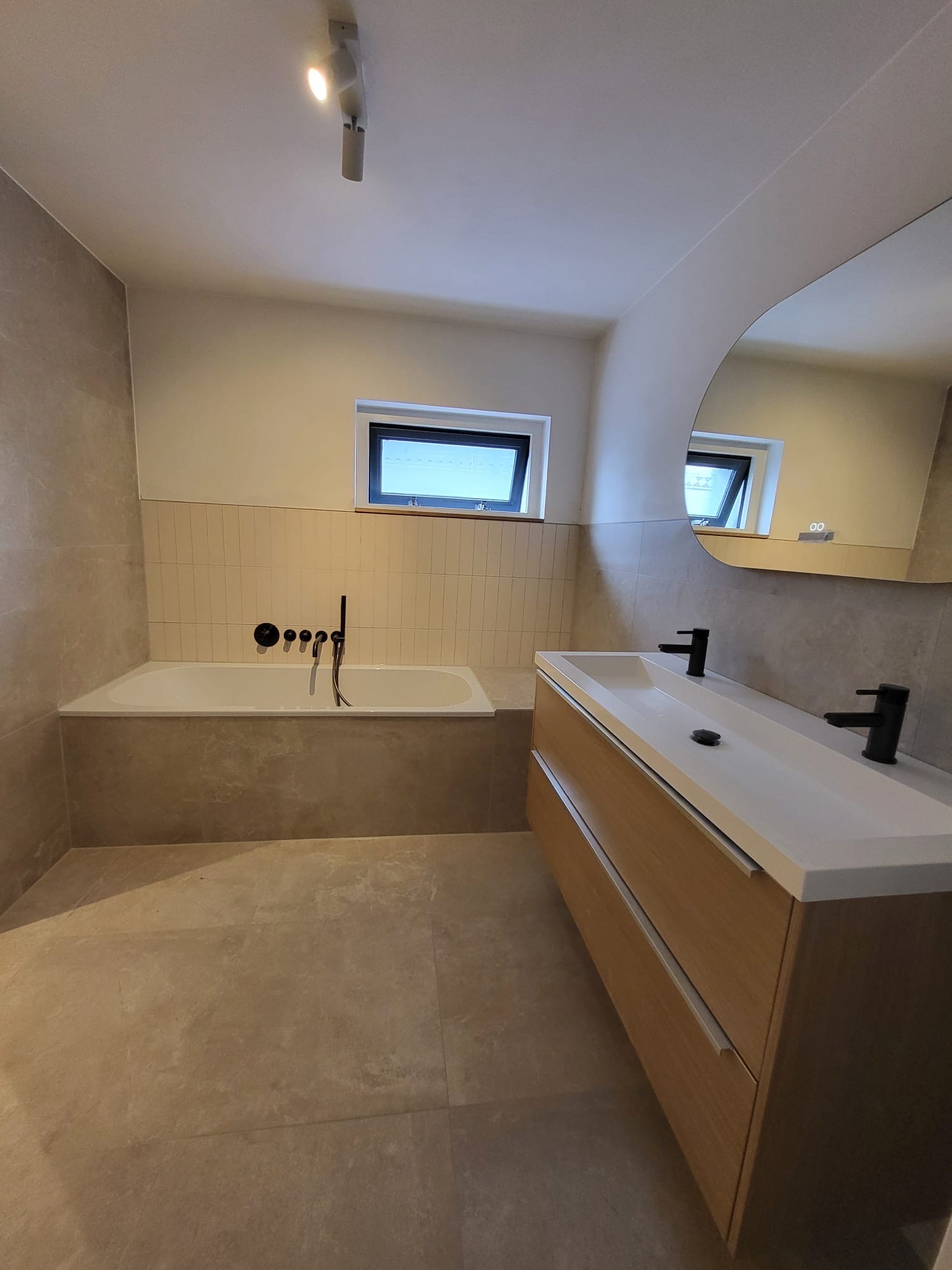 Badkamer met ingebouwd ligbad, grote lichte vloertegels en houten dubbel wastafelmeubel met zwarte kranen