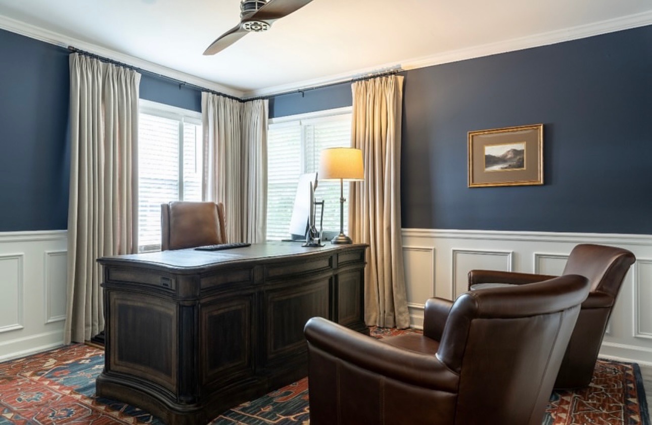 Navy home office with a dark executive desk, leather club chairs, wainscoting, and pleated drapery
