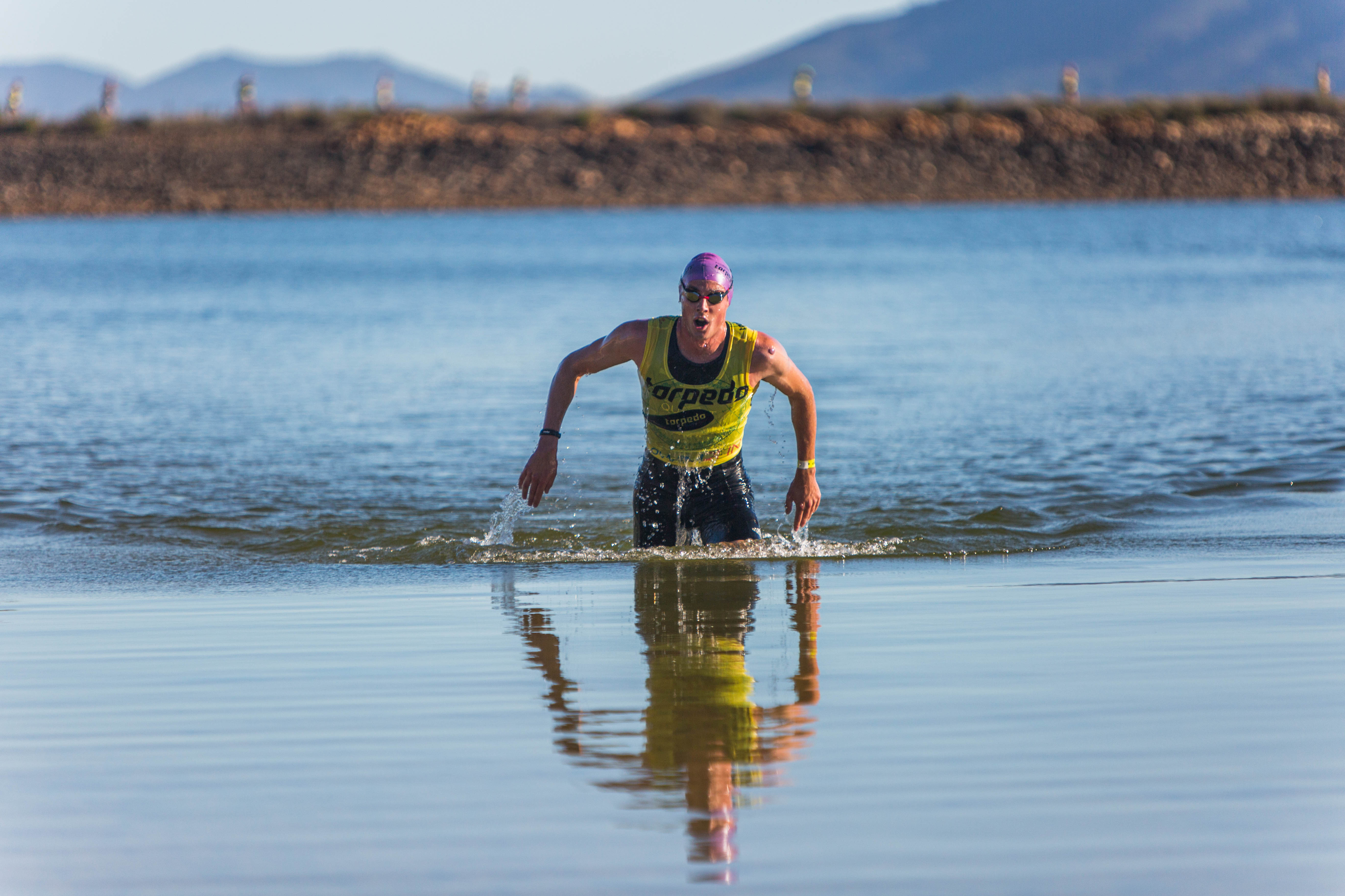 Athlete emerging from calm dam water, reflection visible, mountains behind