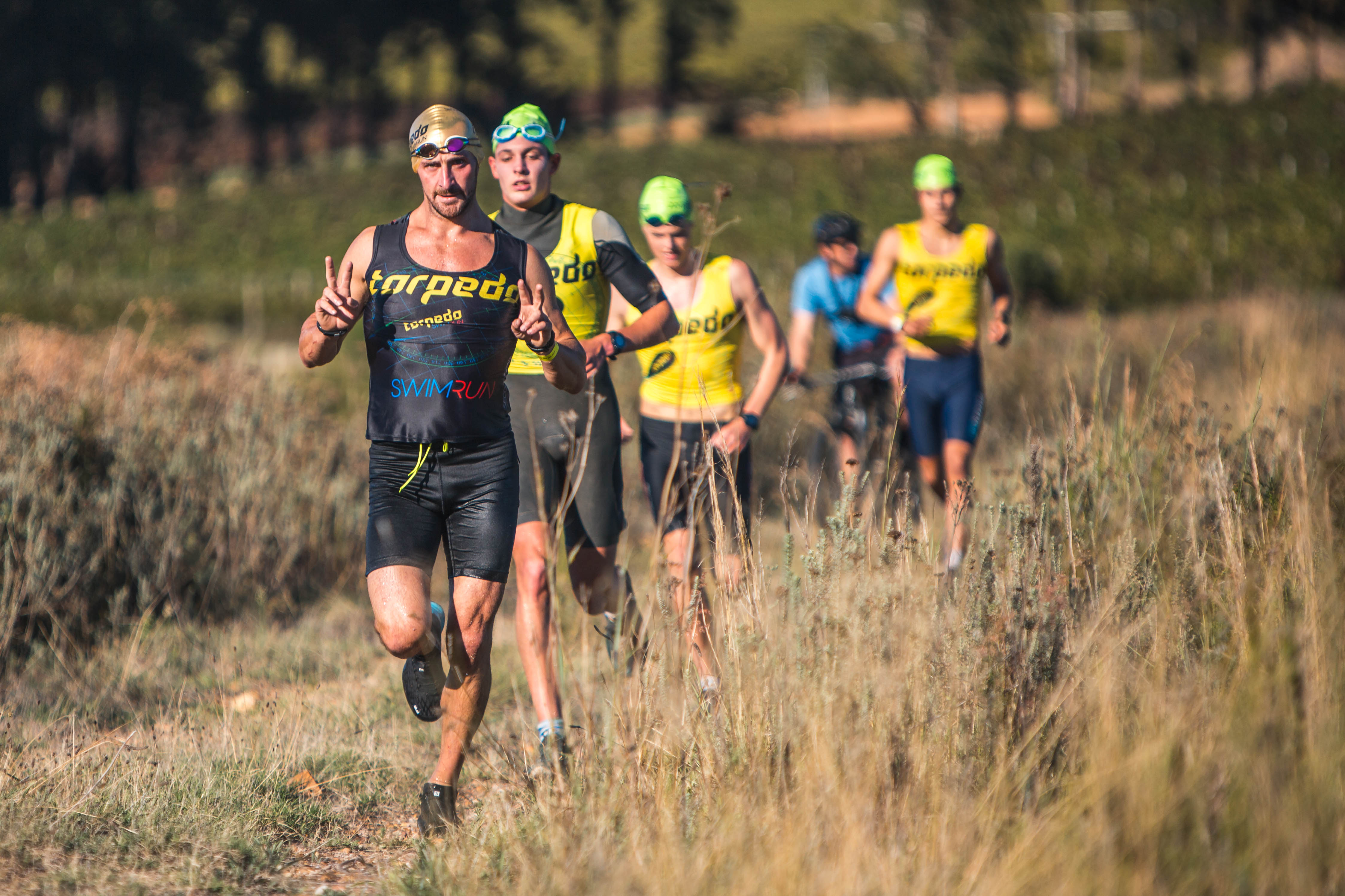 Group of athletes running through dry grassland, leader making peace signs