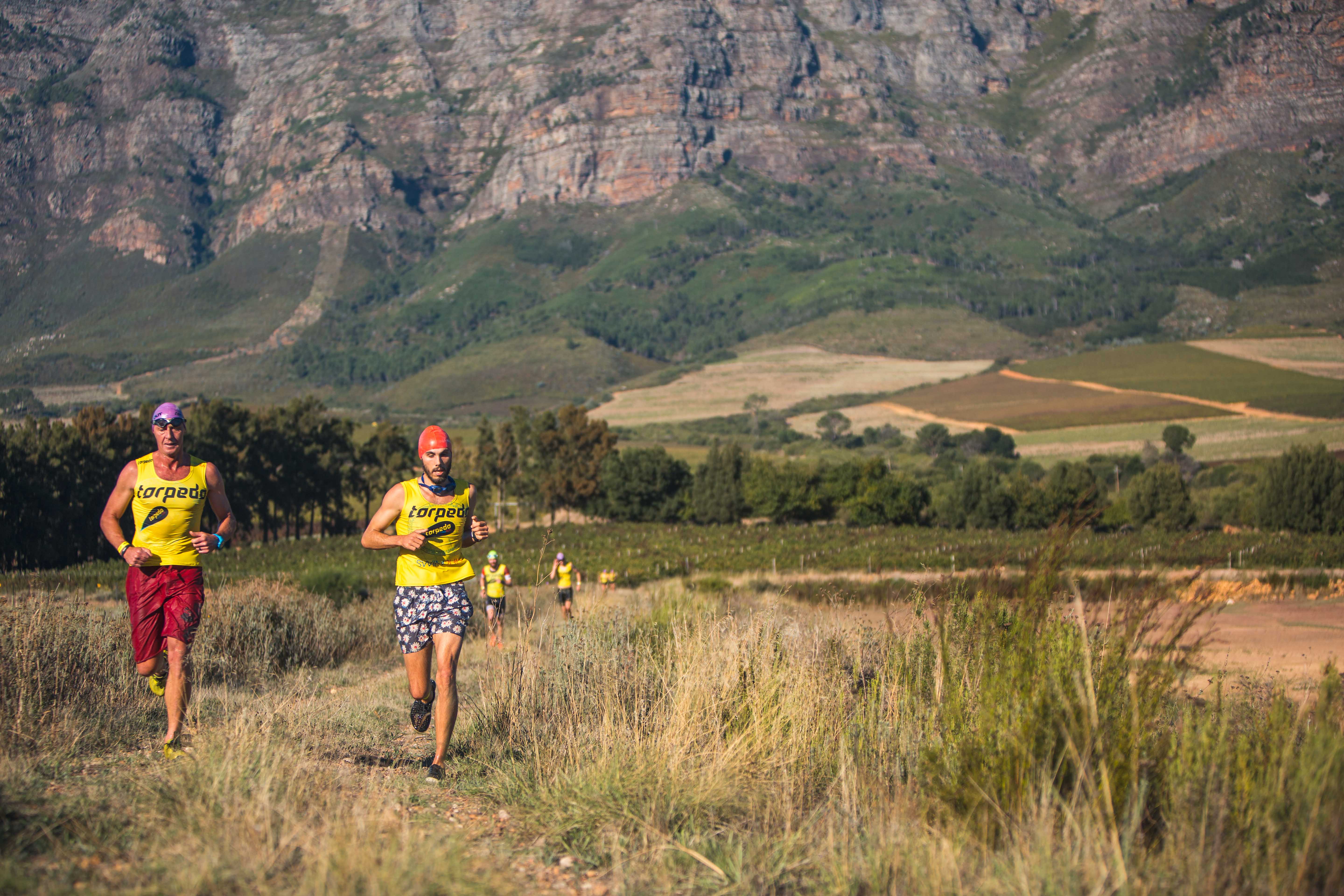 Two athletes running through Winelands fields with mountain backdrop