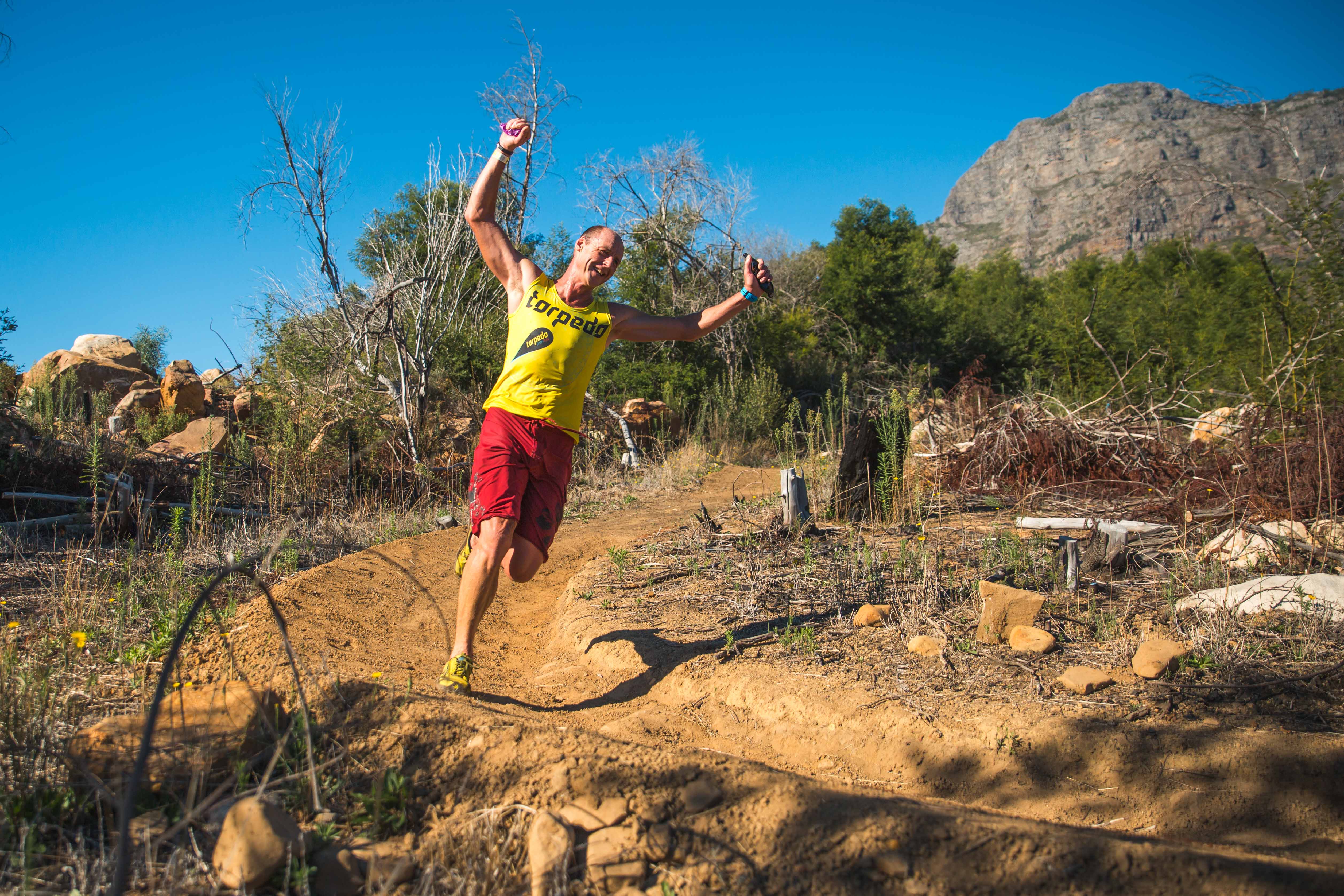 Athlete leaping with arms wide on a dirt trail, mountains behind