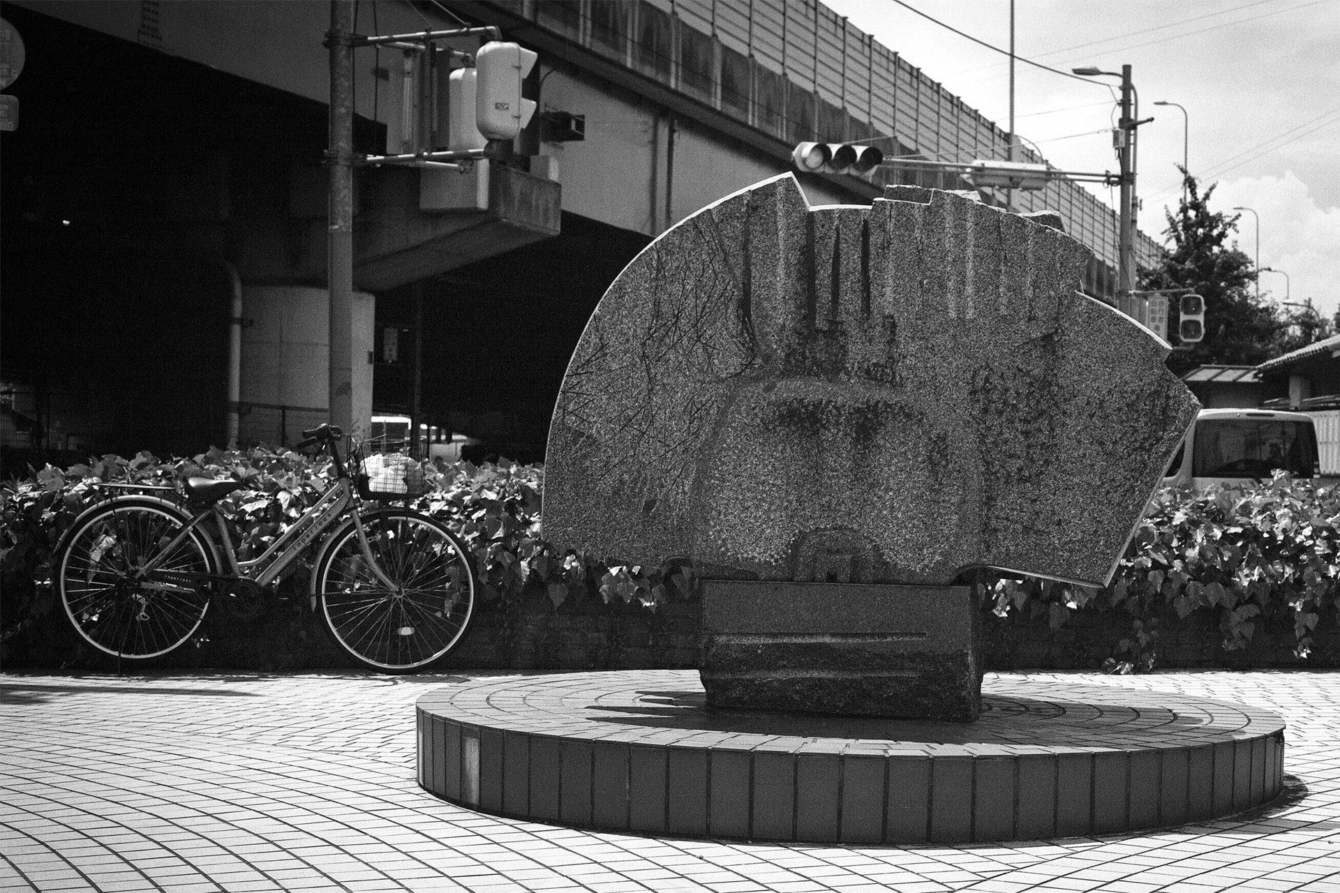 Black and white abstract stone sculpture on circular base with bicycles parked elevated highway background