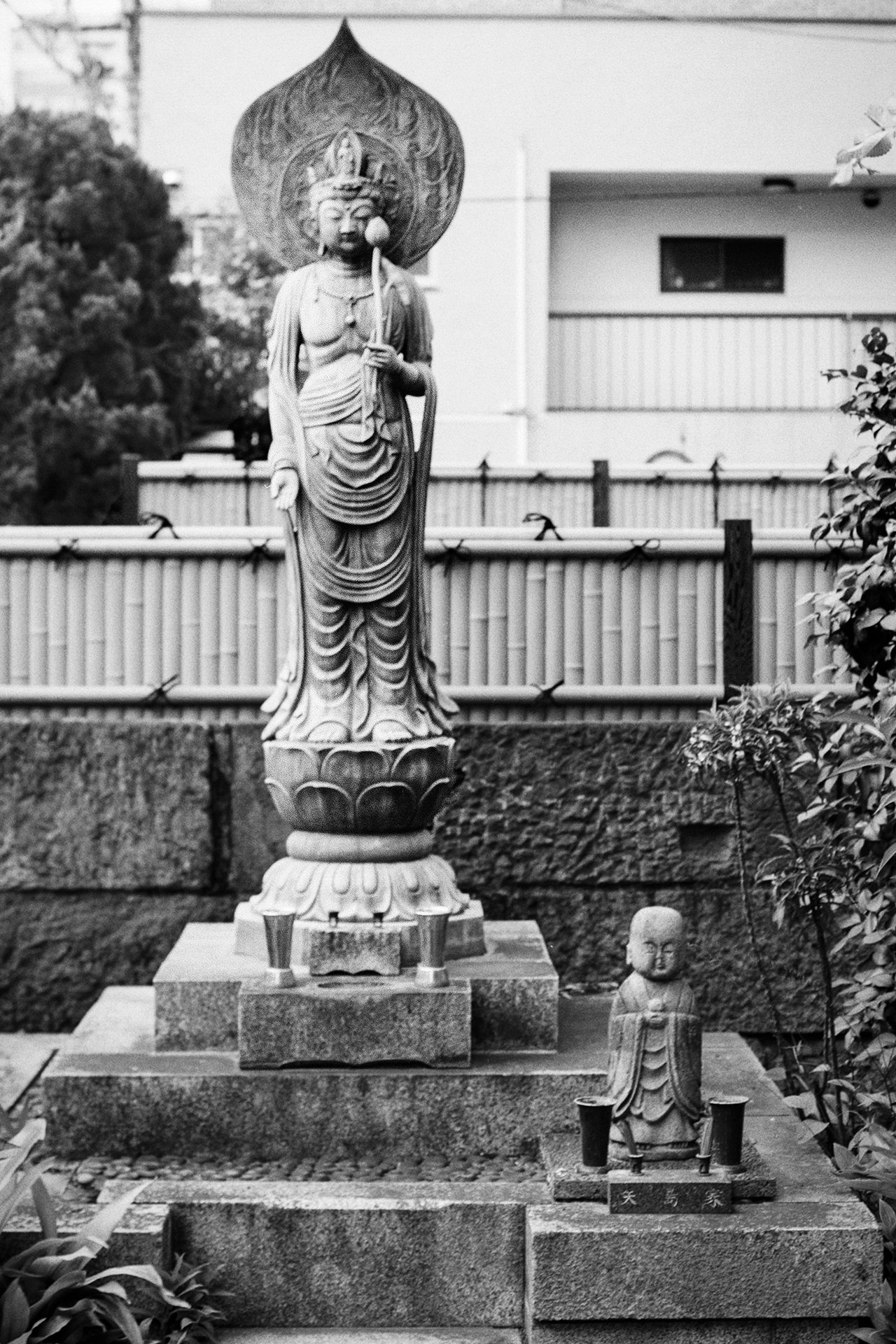 Black and white Buddhist Kannon statue on lotus pedestal with smaller Jizo statue in garden setting