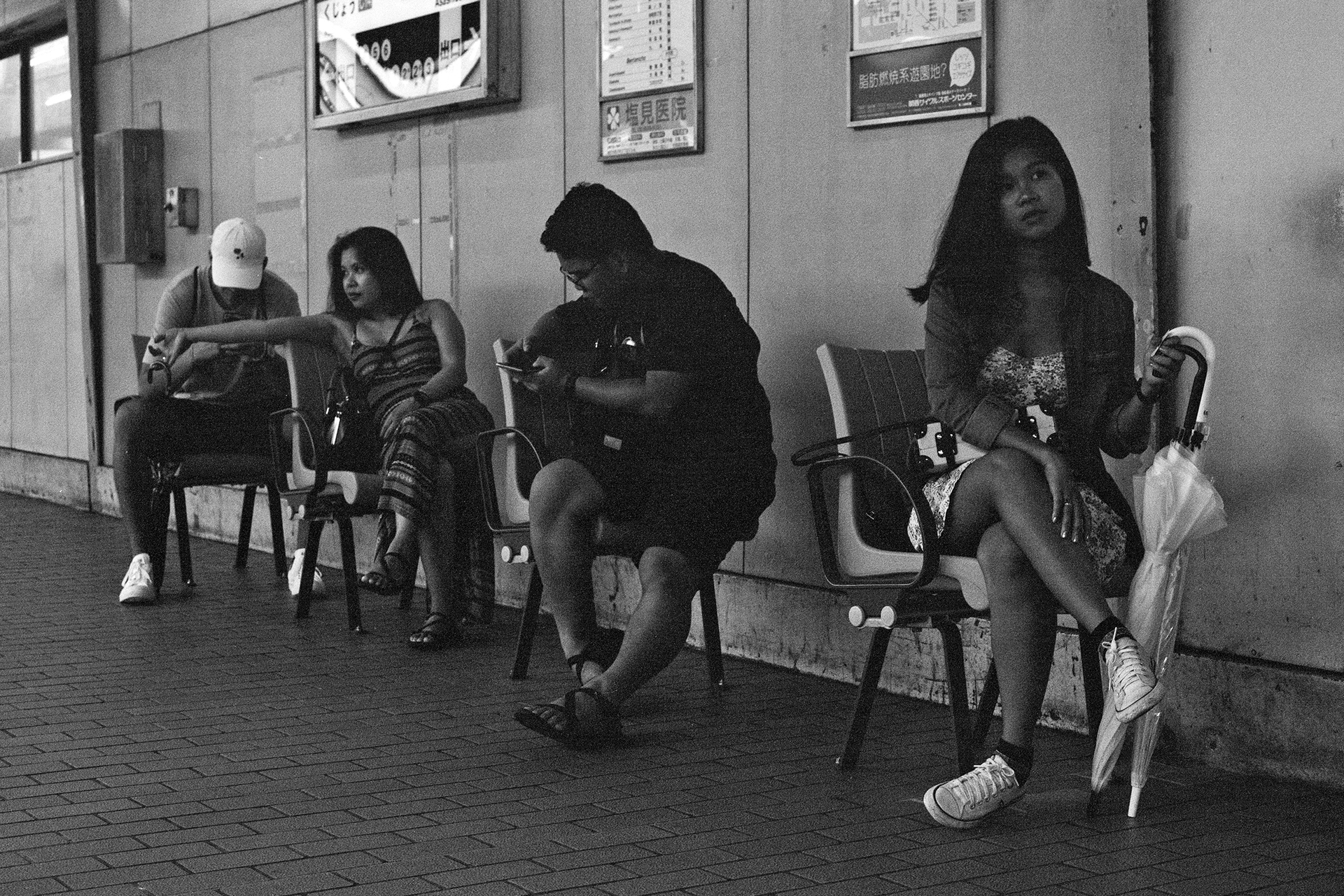 Black and white group of people sitting on benches at station waiting area with Japanese signs