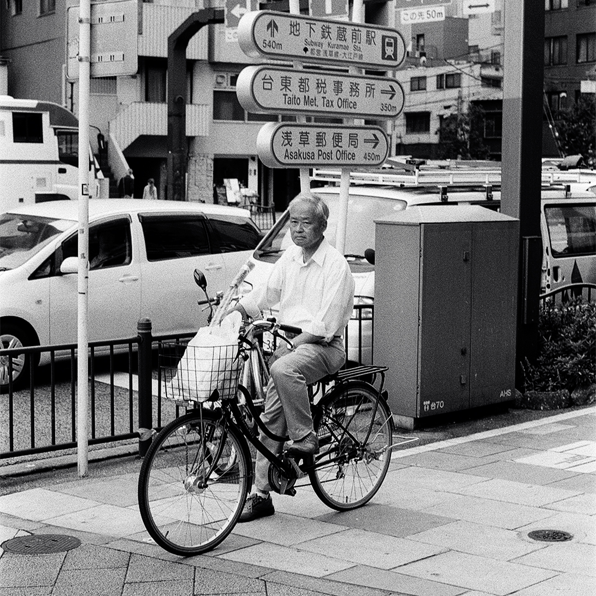 Black and white elderly man on bicycle at intersection with Japanese directional signs Kuramae Station Asakusa Post Office