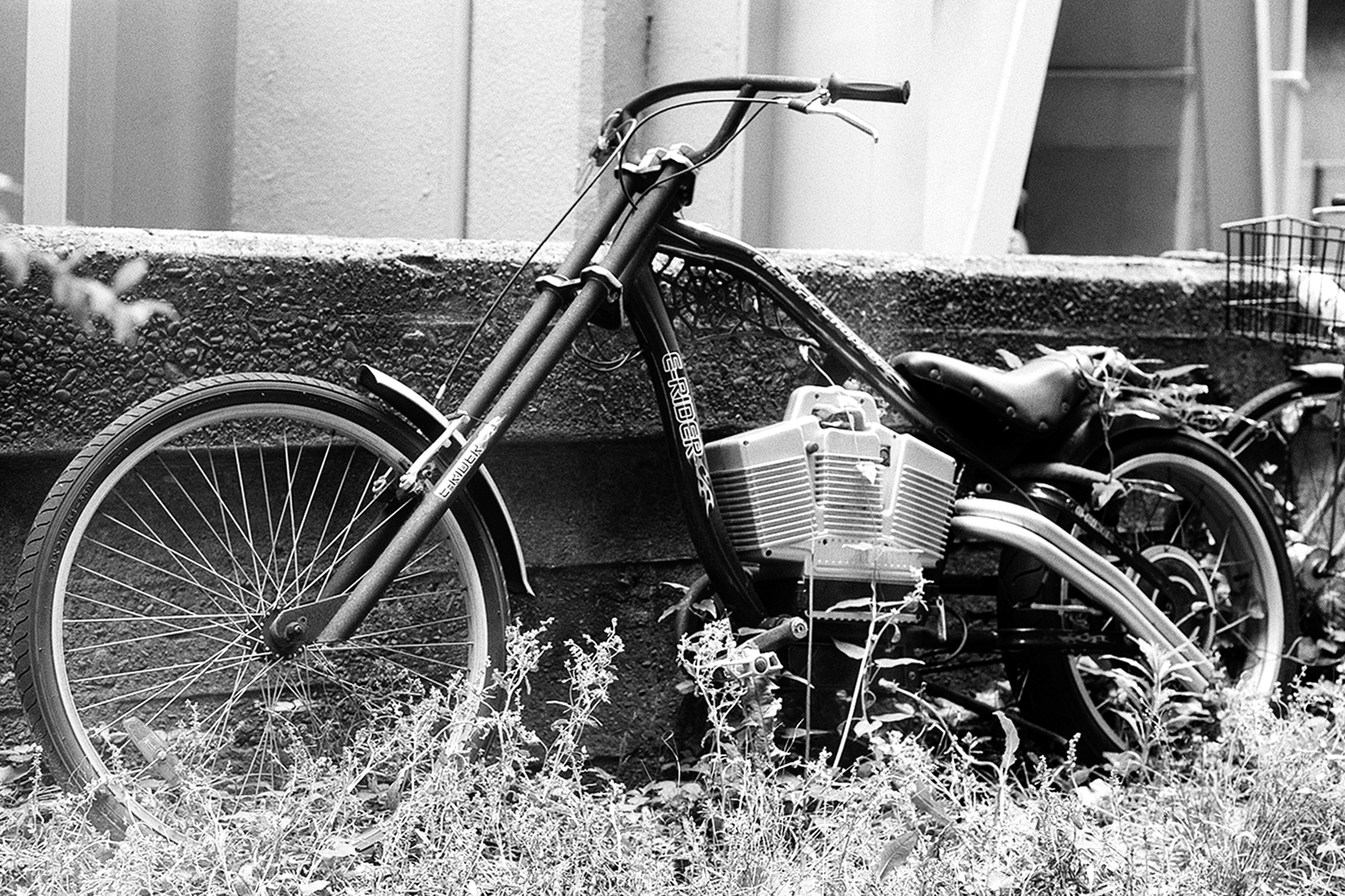 Black and white custom E-Rider motorized bicycle leaning against concrete wall overgrown grass