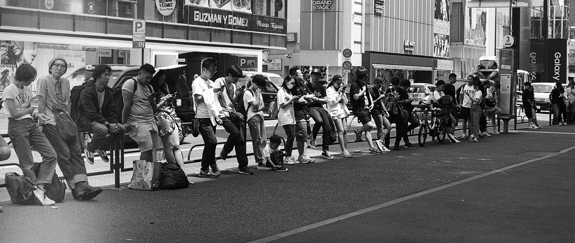 Black and white panoramic Shibuya street crowds sitting along barrier on phones commercial signage