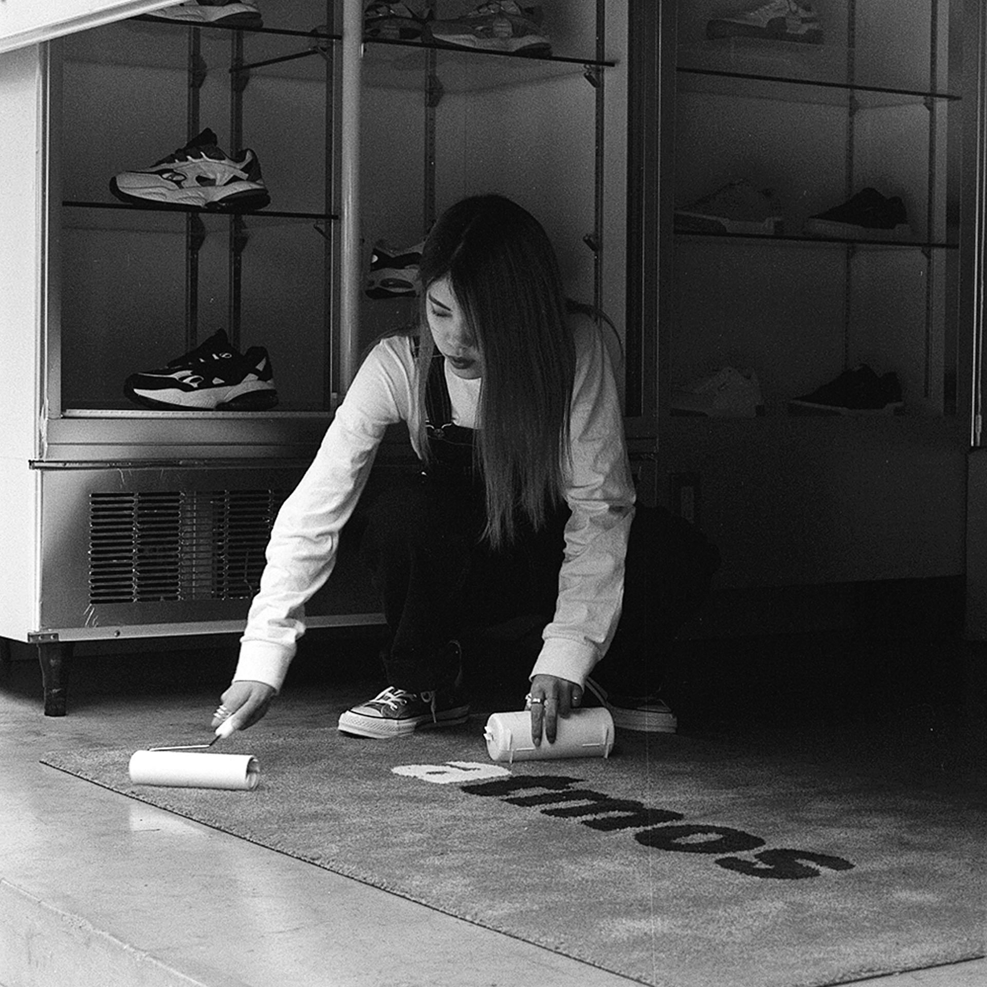Black and white young woman in overalls cleaning floor mat with lint roller sneaker store display