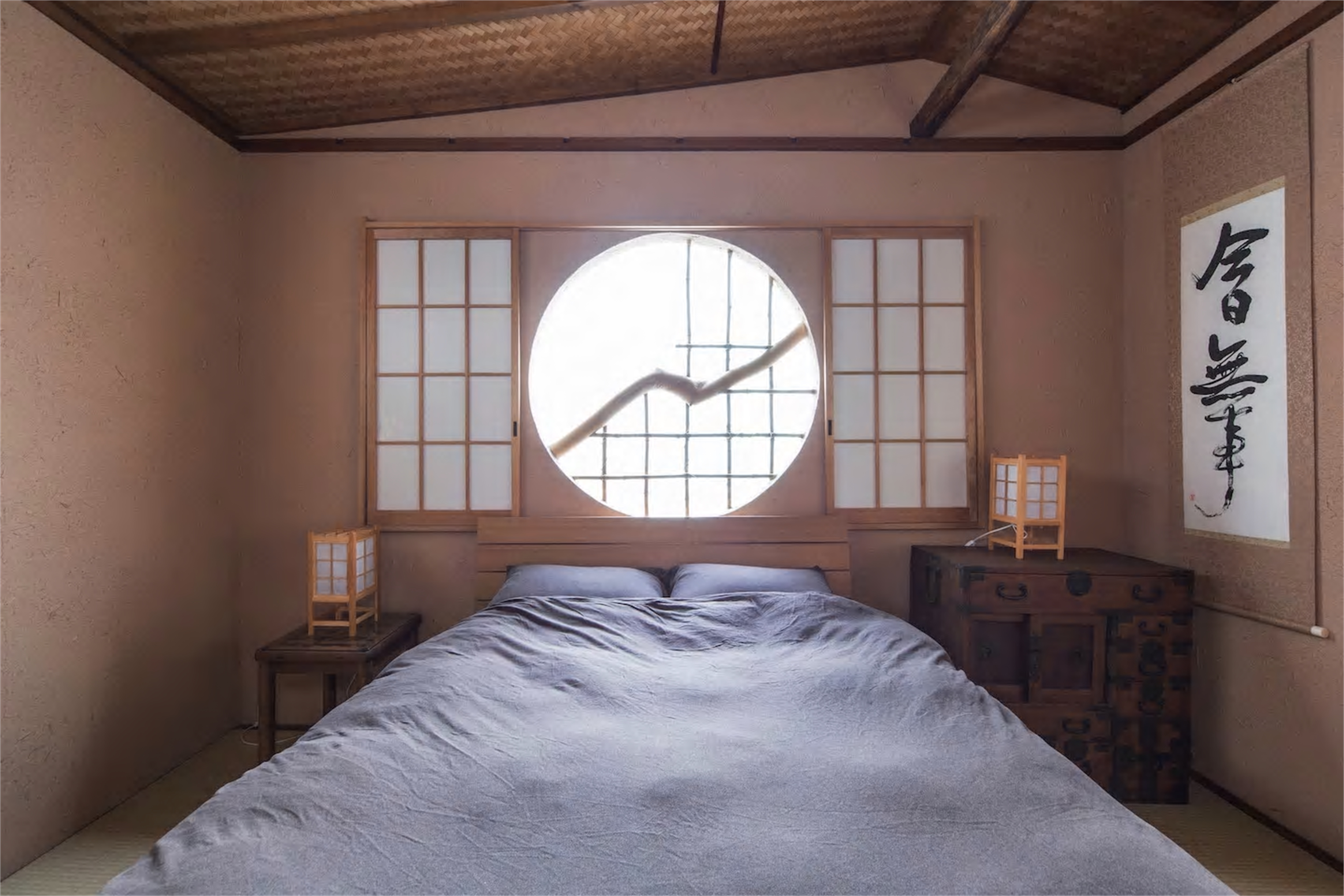 Jizo-An tatami bedroom with double bed in grey-blue linens, circular marumado window framed by shoji screens, woven bamboo ceiling, calligraphy scroll, and antique tansu chest