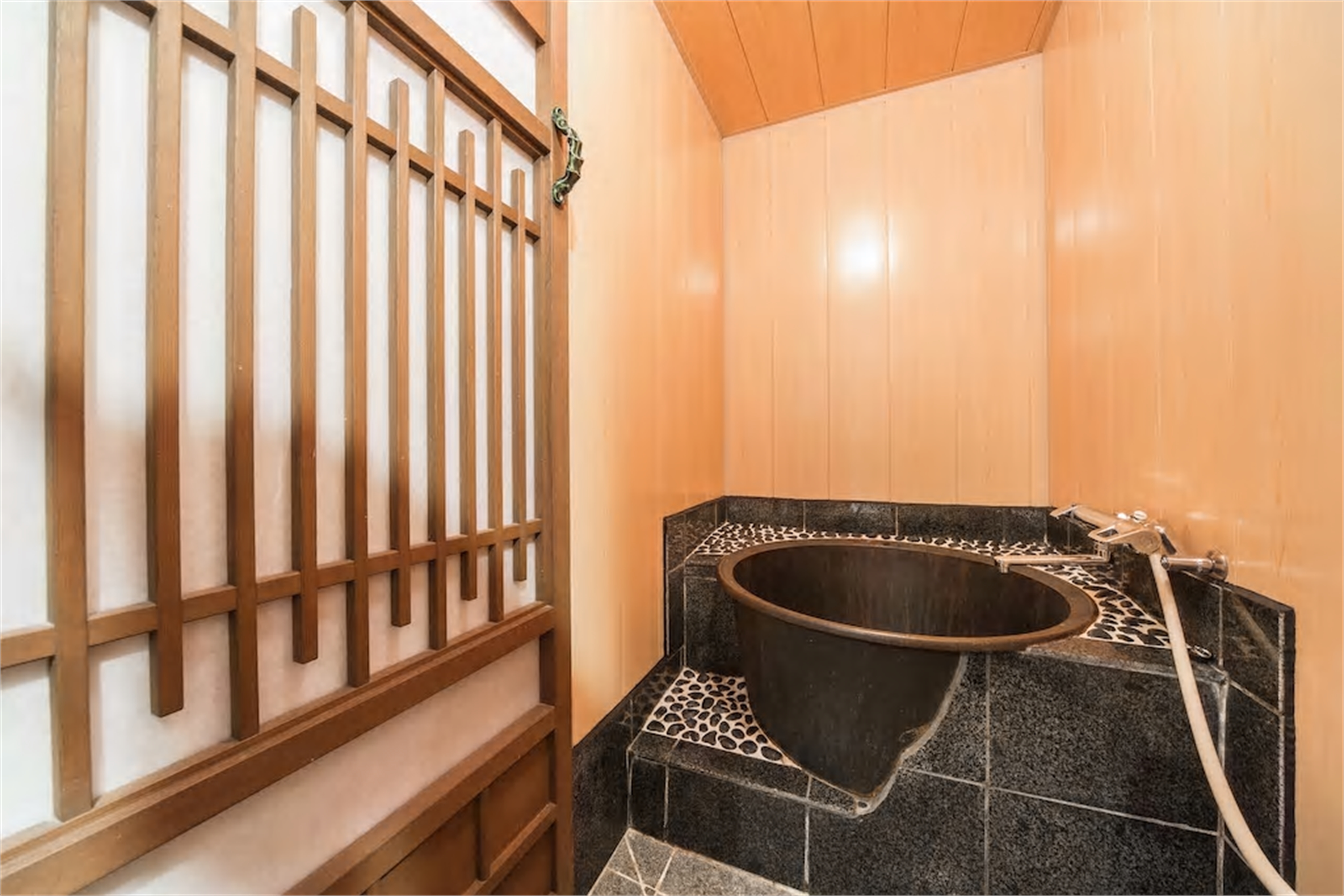 Bathroom with deep black ceramic goemon soaking tub, dark granite tiles, river pebble accents, hinoki wood paneling, and wooden lattice screen