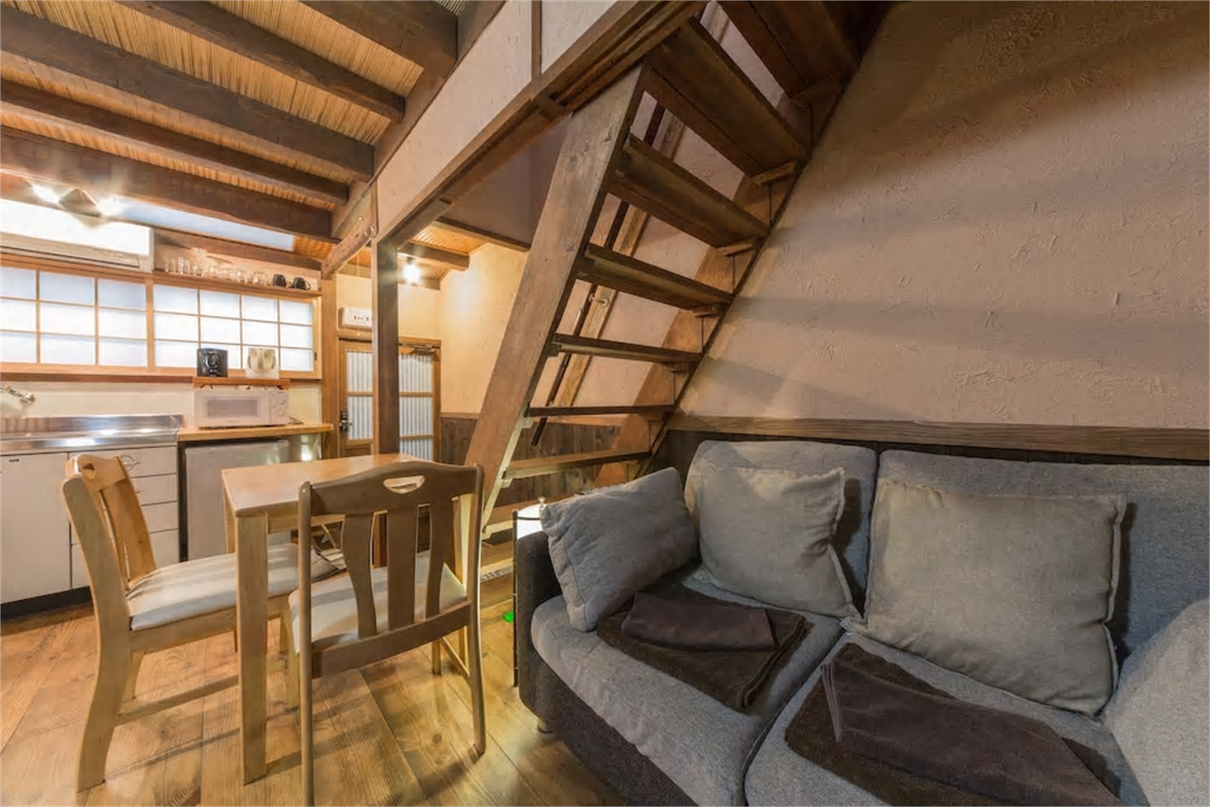 Open-plan living with grey sofa, wooden dining area, steep loft ladder, exposed beams, bamboo reed ceiling, and shoji screen windows