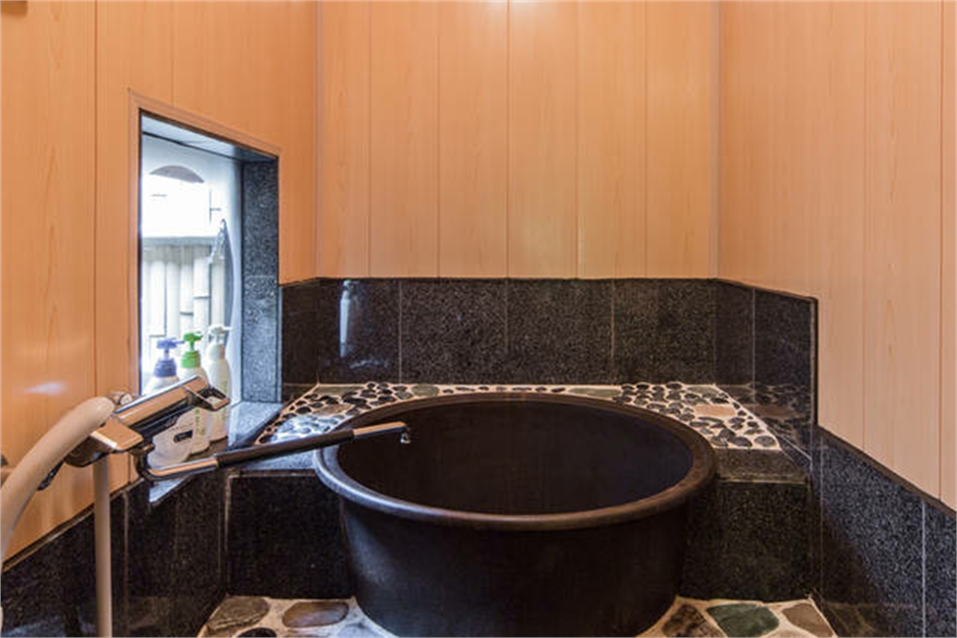 Traditional Japanese bathroom with deep black ceramic goemon-style soaking tub, hinoki wood paneling, dark granite tiles, and river pebble accents