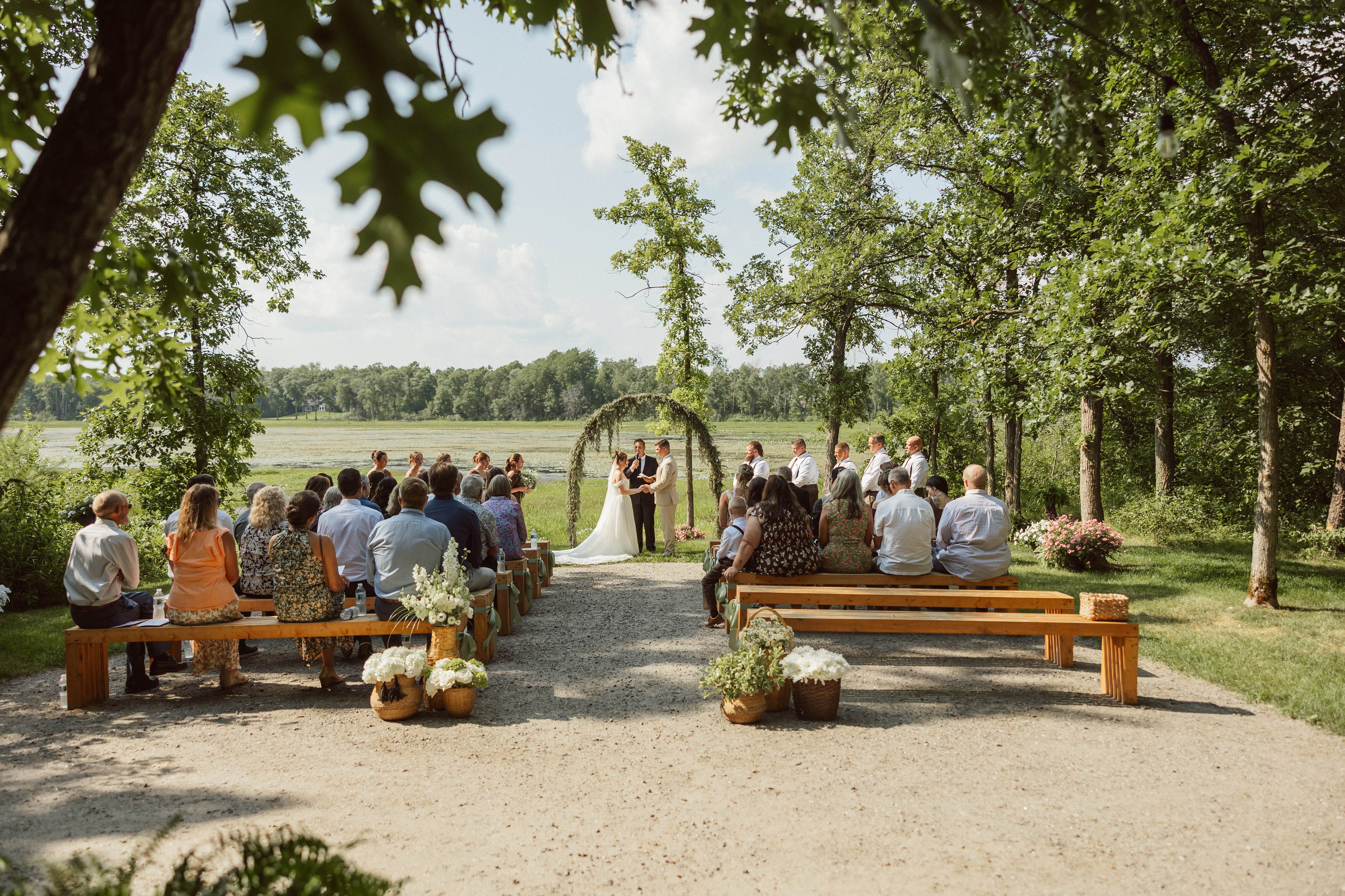 A lakeside wedding ceremony framed by leafy trees with guests gathered along the water