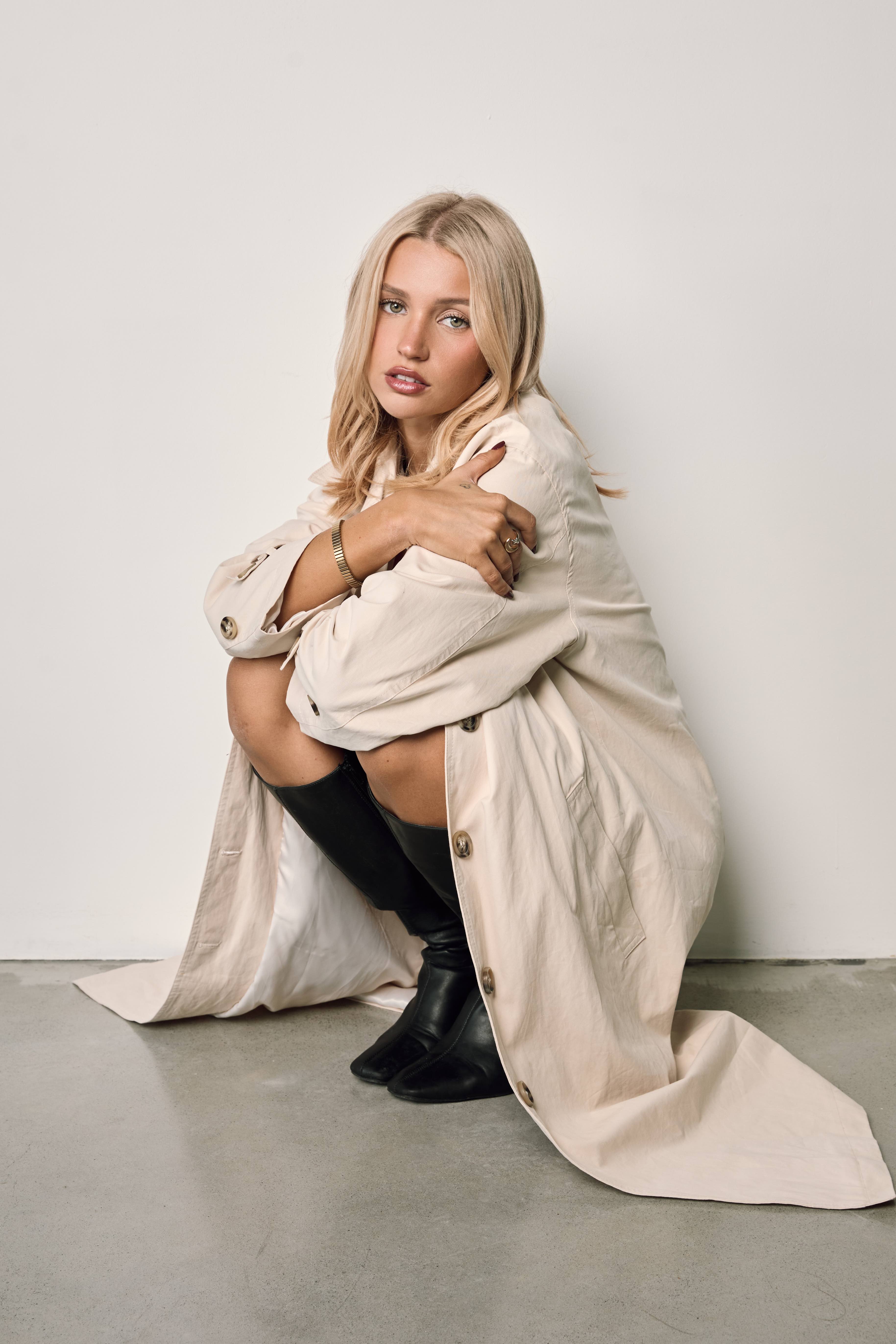 Kyla crouching in a cream trench coat and black knee-high boots