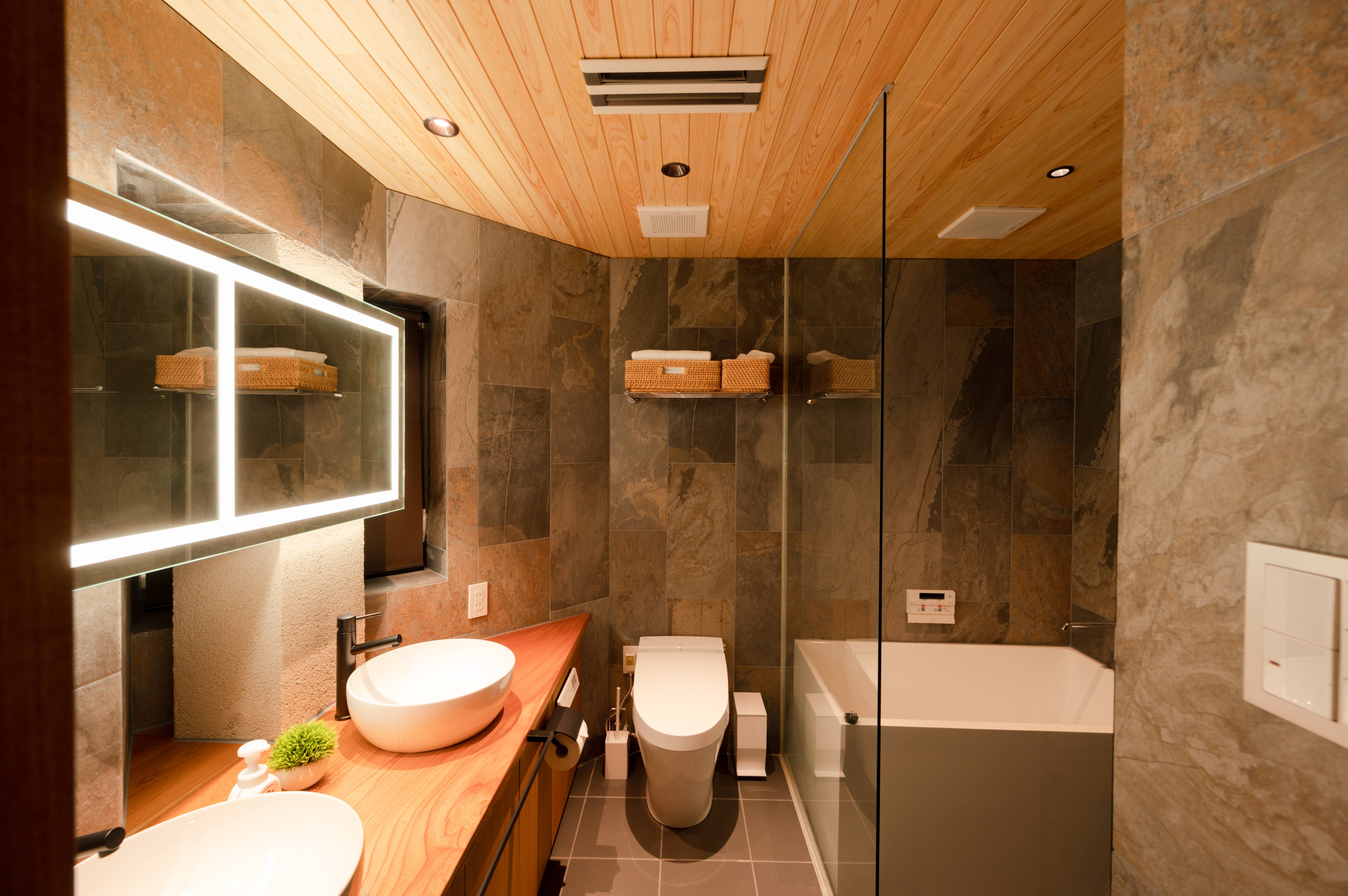 Spa-like bathroom with natural stone walls, light wood ceiling, LED-backlit mirror, dual vessel sinks, and glass-enclosed soaking tub