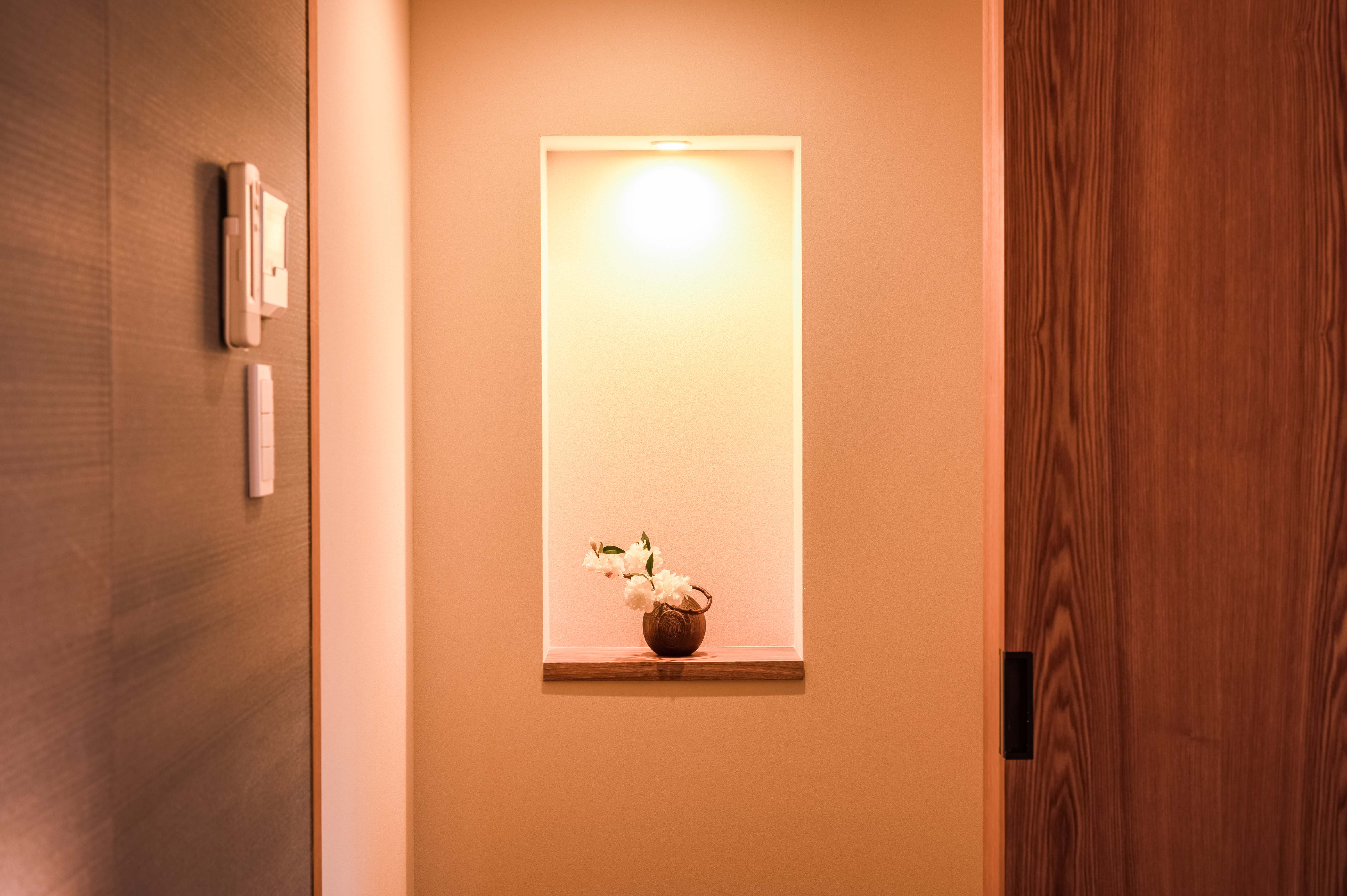 Warm hallway with illuminated recessed niche displaying ceramic vase with white flowers, framed by warm wood paneling