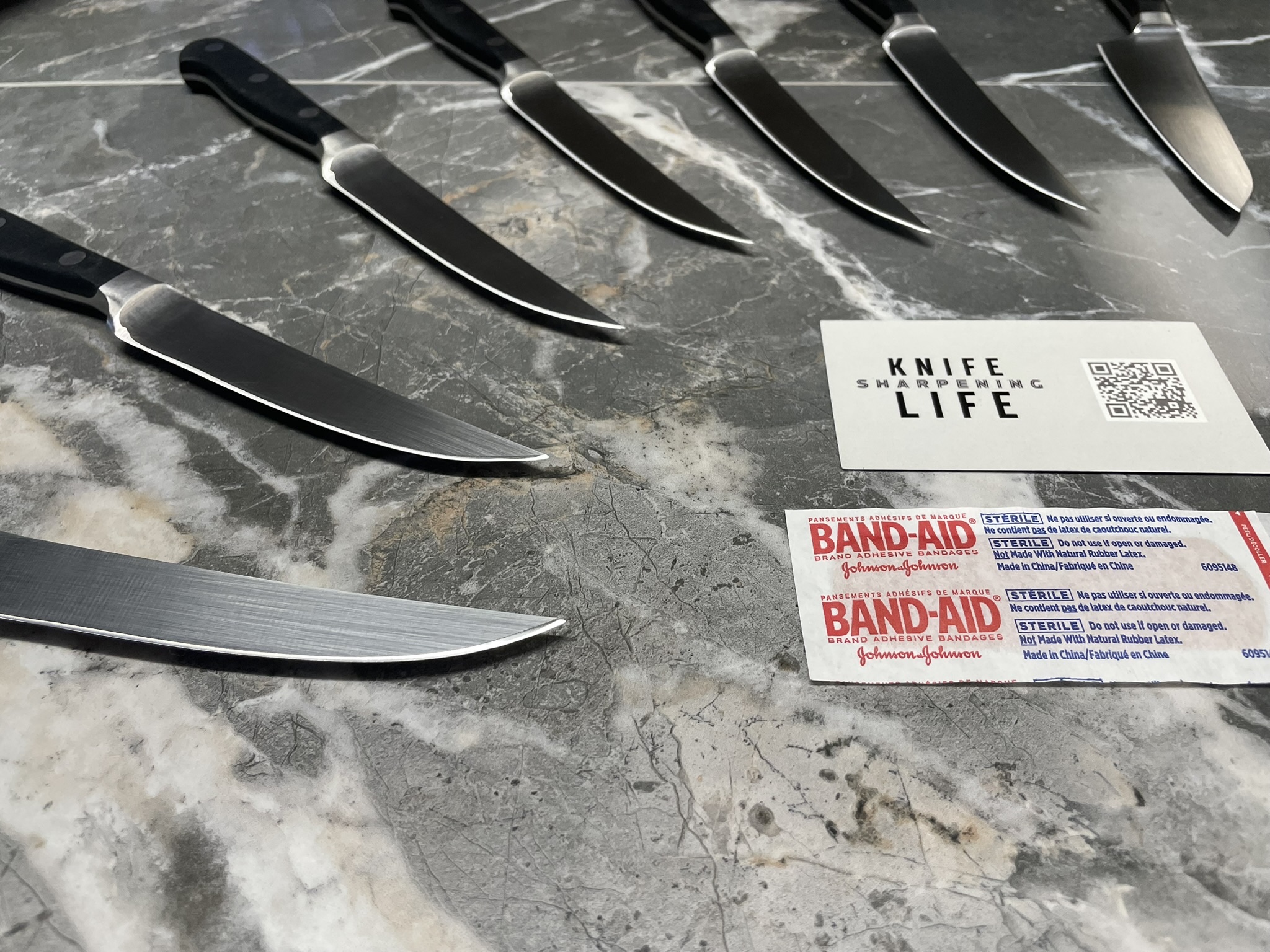Multiple sharpened knives fanned out alongside a Knife Life Sharpening business card on dark marble