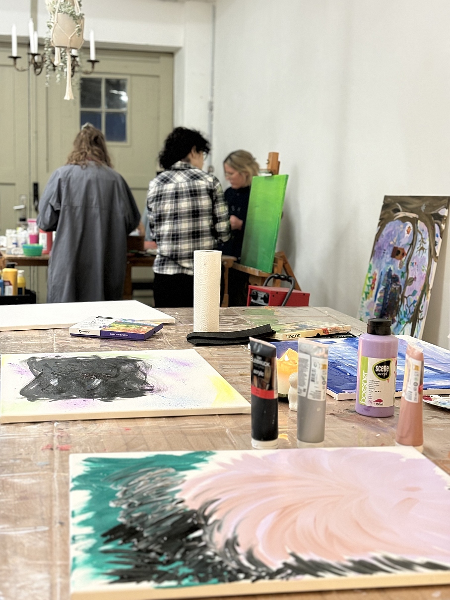 Drei Frauen malen im Kreativraum an Leinwänden, im Vordergrund Acrylfarben und Pastellkreiden auf einem Arbeitstisch